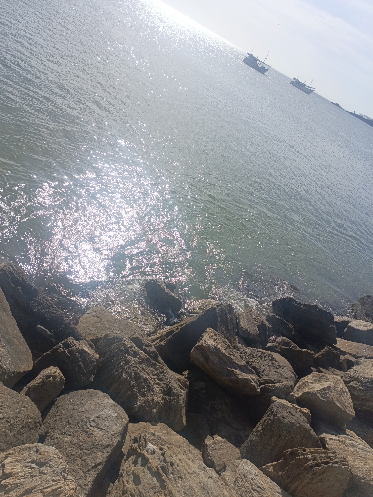 The image shows a rocky shoreline with the ocean in the background, presumably in Juan Griego, Venezuela. Large, angular rocks of varying sizes form a barrier along the edge of the water. The ocean water reflects sunlight, creating a shimmering effect on the surface. In the distance, there are a few boats visible on the water. The sky is light, suggesting it is daytime. The overall scene captures a coastal environment with a rocky breakwater.