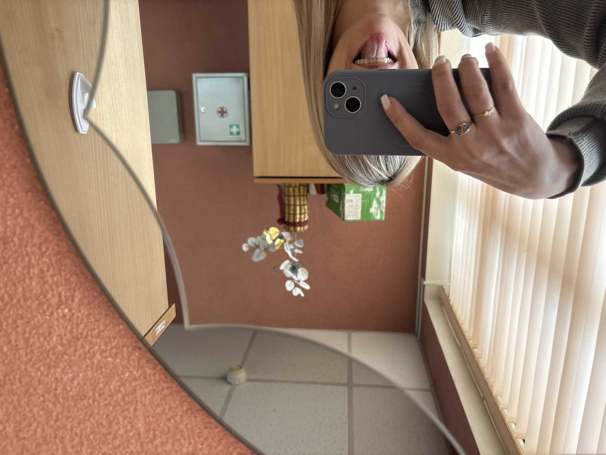 A mirrored selfie shows a woman holding a gray phone. She's sticking her tongue out. The reflection reveals a bathroom setting. A first aid kit is mounted on the wall. A wooden cabinet is also visible. A decorative floral arrangement and striped blinds are in the scene. The mirror is attached to a wall covered in an orange, textured material. The floor is tiled. There is an object on the floor beneath the flower arrangement, likely a part of it. The scene seems to be taking place in a bathroom in Chaikovsky, Russia, as the location is provided.