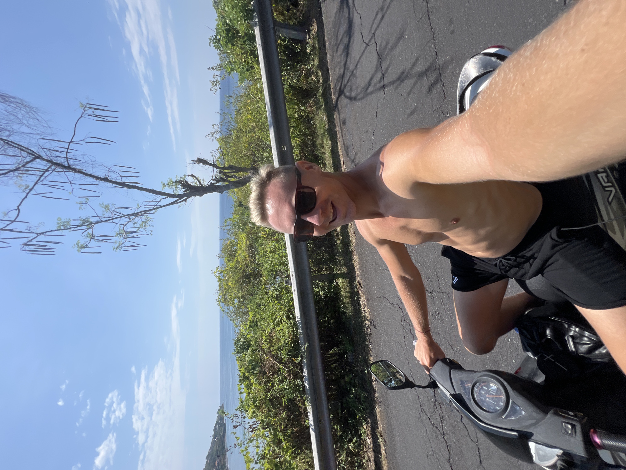 A shirtless man with blonde hair, wearing sunglasses and black shorts, rides a black scooter on a paved road. He is holding the handlebars with his right hand and smiling at the camera, which appears to be a selfie. The scooter's speedometer is visible. In the background, lush green vegetation lines the road, with a metal guardrail separating it from the ocean. A tree with long, dangling seed pods is in the middle ground. The sky is clear blue with scattered white clouds. The location is identified as Abang, Indonesia, likely a coastal road.