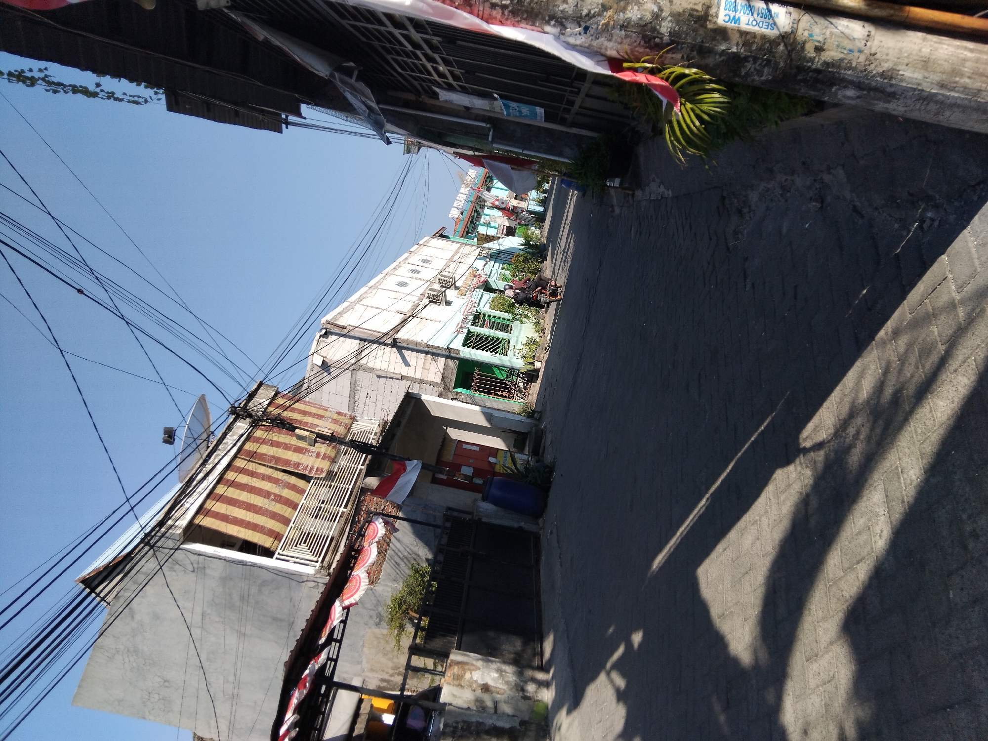 This is a daytime, medium-angle, eye-level shot of a narrow, paved street in the city of Kepuh Kiriman, Indonesia, with buildings on both sides and a clear blue sky above. The street is paved with interlocking dark gray bricks. On the left side of the street, a tall, light gray building with a white balcony railing and red and tan striped awnings is visible. A black metal gate with a concrete wall is in front of this building, and a large blue plastic barrel is placed near the gate. An Indonesian flag is mounted on the side of this building. Further down the street on the left, a multi-story building with a light blue facade and white window frames is visible, with air conditioning units mounted on the exterior.
On the right side of the street, a dark-colored building with a corrugated metal roof is present. The front of this building features a metal gate with vertical bars, and various items are hung on a pole in the foreground, including stickers and what appears to be a partial Indonesian flag. Several motorcycles are present on the street, with two people on one motorcycle riding away from the camera. Other motorcycles are parked along the sides of the street. Plants and greenery are visible in pots and growing along the edges of the buildings and fences. Electrical wires are strung across the sky, obscuring parts of the view. Shadows from overhead structures are cast across the street. The overall impression is of a densely built urban environment.