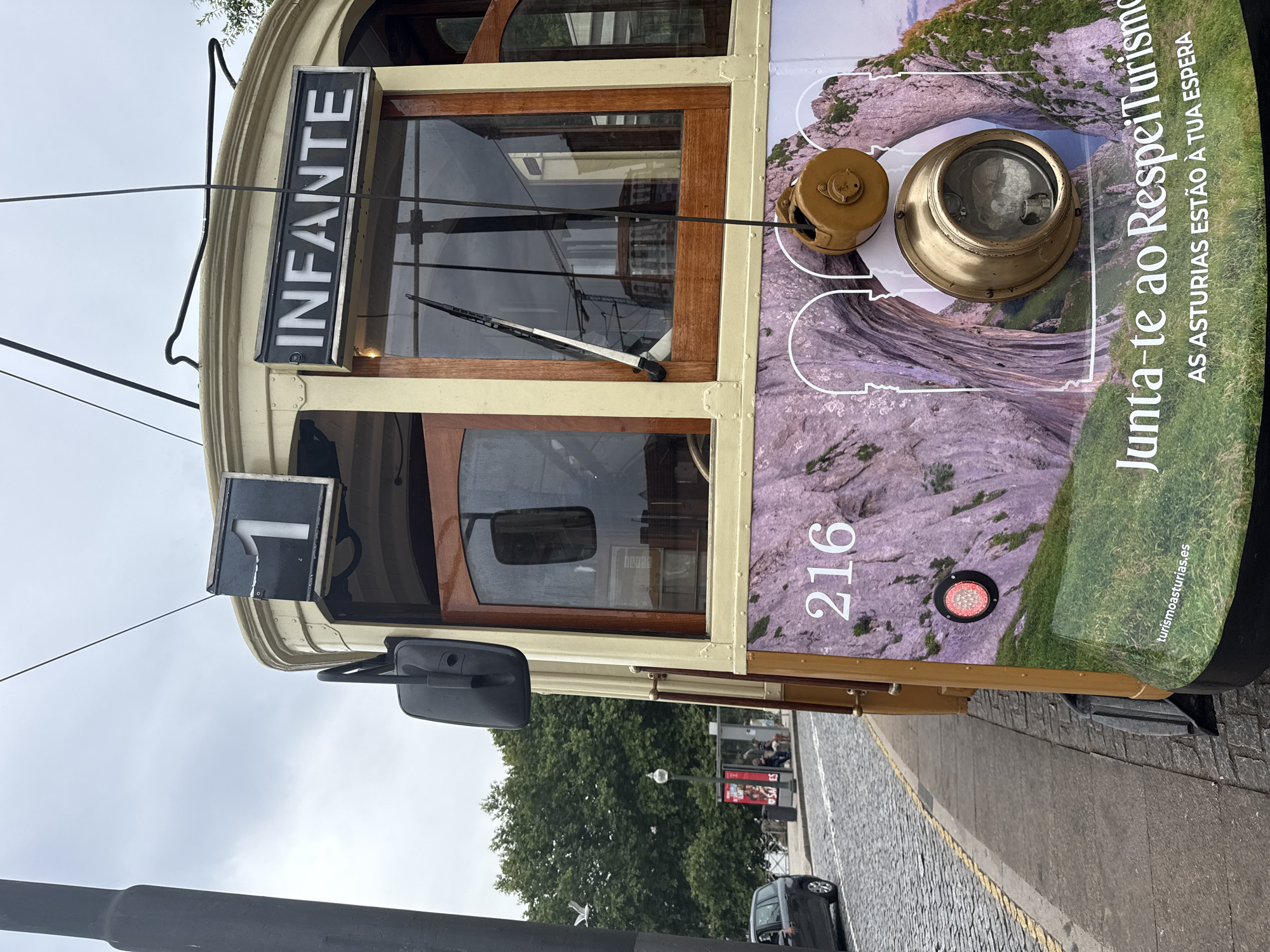 The image shows a vintage tram in Porto, Portugal, on a cloudy day. The tram is cream-colored with wooden accents and features the number "1" and the word "INFANTE" displayed on its front. The number "216" is visible on the side of the tram, along with a graphic of a mountainous landscape and the text "Junta-te ao RespeitTurismo AS ASTURIAS ESTÃO À TUA ESPERA". A black car is parked on the cobblestone street to the left, and a lamppost and bus stop are visible in the background. Trees and buildings line the street, suggesting an urban setting.