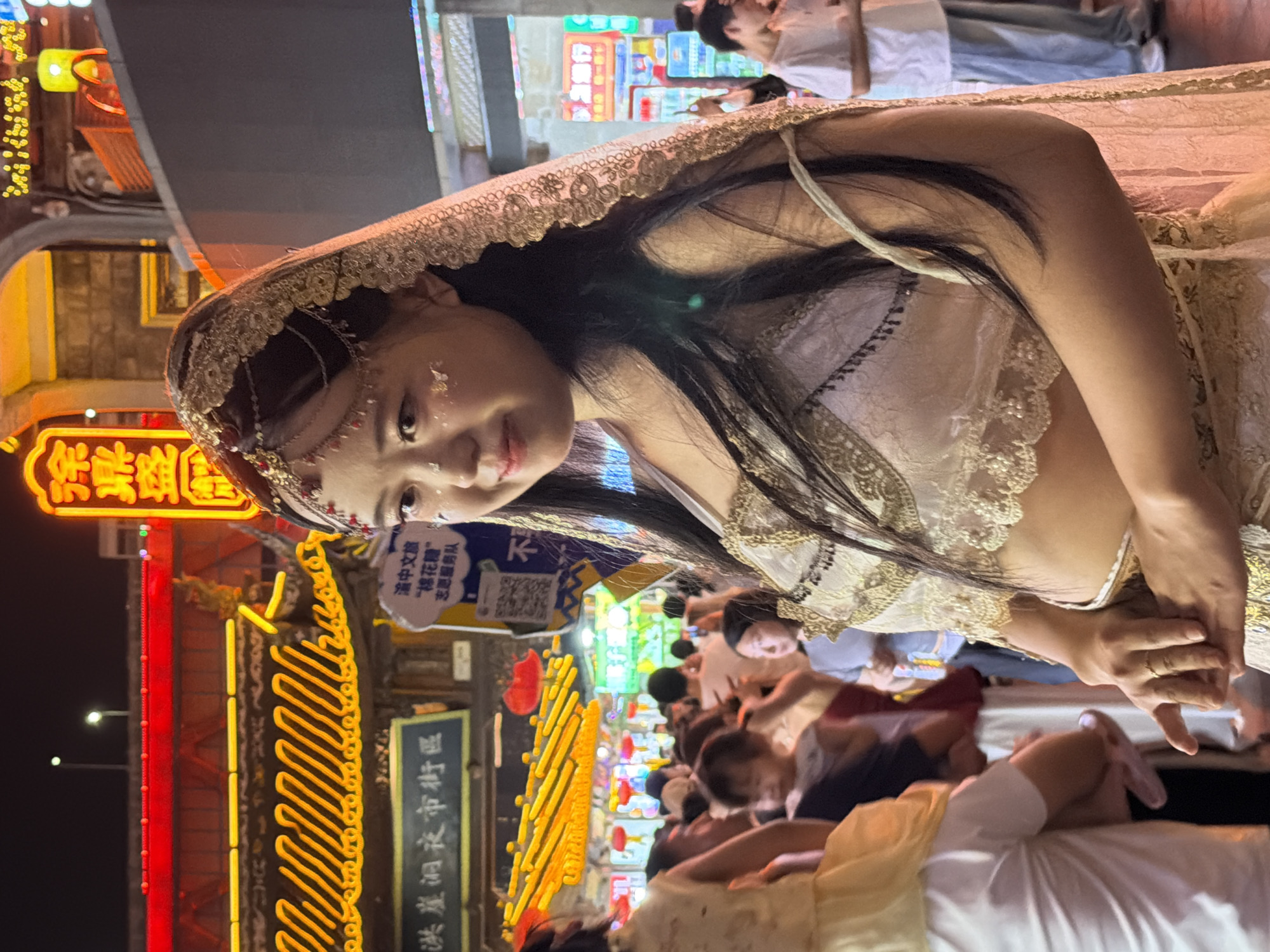 This media file shows a young woman dressed in traditional Indian attire, possibly a bridal outfit, with a gold-colored veil adorned with intricate patterns. She has delicate makeup and a piece of jewelry on her forehead. She is looking slightly downwards and to the side with a soft expression. The background suggests a lively night market scene.
The scene is dimly lit, indicating it is nighttime. There are bright neon signs and string lights contributing to the ambient light. In the background, a crowd of people can be seen, some appearing to be tourists or shoppers enjoying the atmosphere. The woman's attire stands out amidst the more casual clothing of the people around her, suggesting she might be participating in an event or performance.
Visible text in the image includes Chinese characters on a blue and white sign, which appears to be a guide for "Yuzhong District Night Market" and mentions "Yuzhong Chinese Culture and Tourism" and "Cotton Candy Volunteer Service Team." Another neon sign in the background has Chinese characters in bright yellow. The overall setting is a bustling street at night, filled with the vibrant energy of a market. The woman's posture and gaze create a sense of gentle curiosity or engagement with her surroundings.