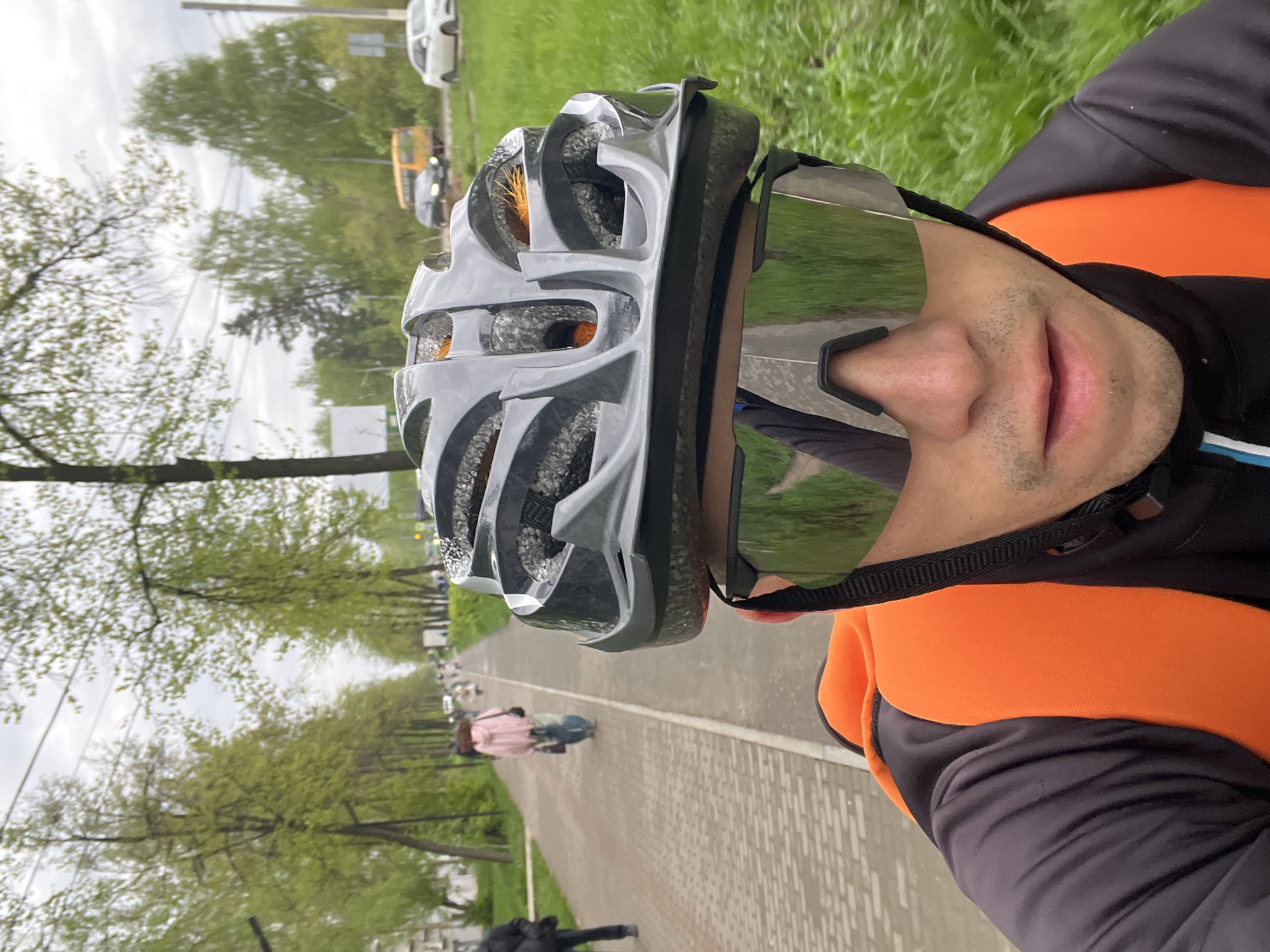 A cyclist wearing a gray helmet and reflective sunglasses is seen from a first-person perspective on a paved path. The path is surrounded by green trees, suggesting a park or green area. In the background, there's a glimpse of traffic, including a yellow bus and white cars, indicating the location is near a road or street. Other cyclists and pedestrians can be seen further down the path. Given the specification of Chaikovsky, Russia, this likely represents a public green space within the city. The cyclist is wearing a black and orange jacket. The ground is constructed of rectangular paving stones.