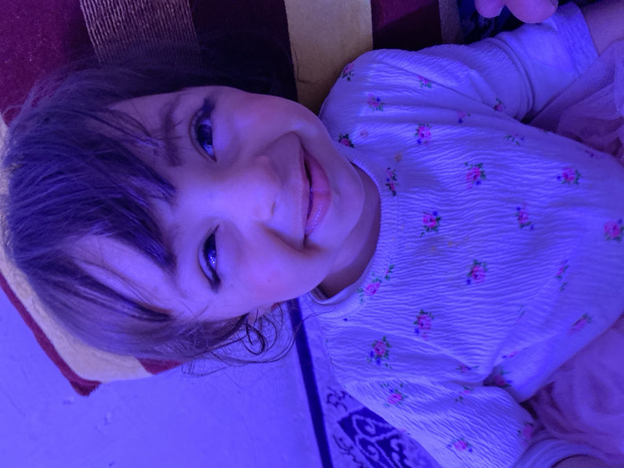 An indoor close-up photograph features a young child, likely a girl, smiling broadly and playfully looking directly into the camera. The image is characterized by a dominant purple-blue artificial light source that casts a strong hue over the entire scene, affecting skin tones and object colors.
The child is positioned diagonally across the frame, with her head tilted to the right. She has dark hair with bangs falling over her forehead, and her eyes, appearing dark blue due to the lighting, are wide open and sparkling. Her cheeks are slightly flushed, and her mouth is open in a wide smile, with her tongue peeking out. The expression conveys joy and a sense of mischief or playfulness.
She is wearing a long-sleeved, light-colored top that has a textured, slightly crinkled fabric. The top is adorned with small, delicate floral patterns in what appear to be muted pink, green, and possibly yellow colors, though these colors are heavily influenced by the purple-blue lighting. On the right side of the frame, there is a hint of light pink or lavender frilly material, possibly part of a skirt or dress, adding to the soft aesthetic.
The background shows elements of an indoor setting. To the left and behind the child's head, there is a soft furnishing with distinct reddish-brown and beige stripes, likely a cushion, pillow, or part of a sofa. On the bottom right, a segment of a darker, possibly deep blue or purple, patterned fabric is visible, suggesting a rug or another piece of decorative textile. The overall lighting suggests an evening or nighttime setting where specific mood lighting is in use. No discernible text is visible in the image.