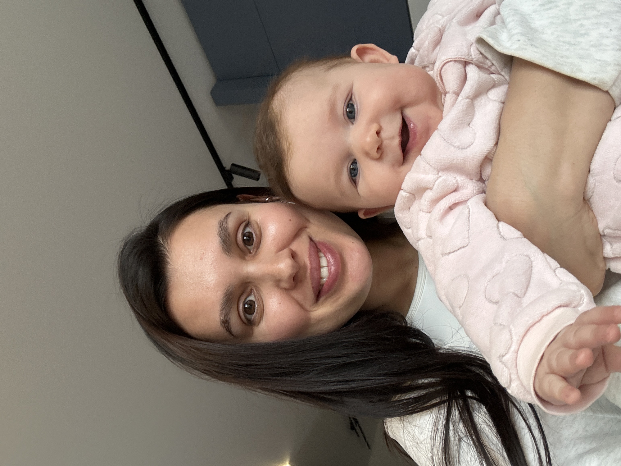 A woman with dark hair holds a baby wearing a light pink onesie with hearts. The baby has blue eyes and is smiling. The location is not discernible as Chaikovsky, Russia.