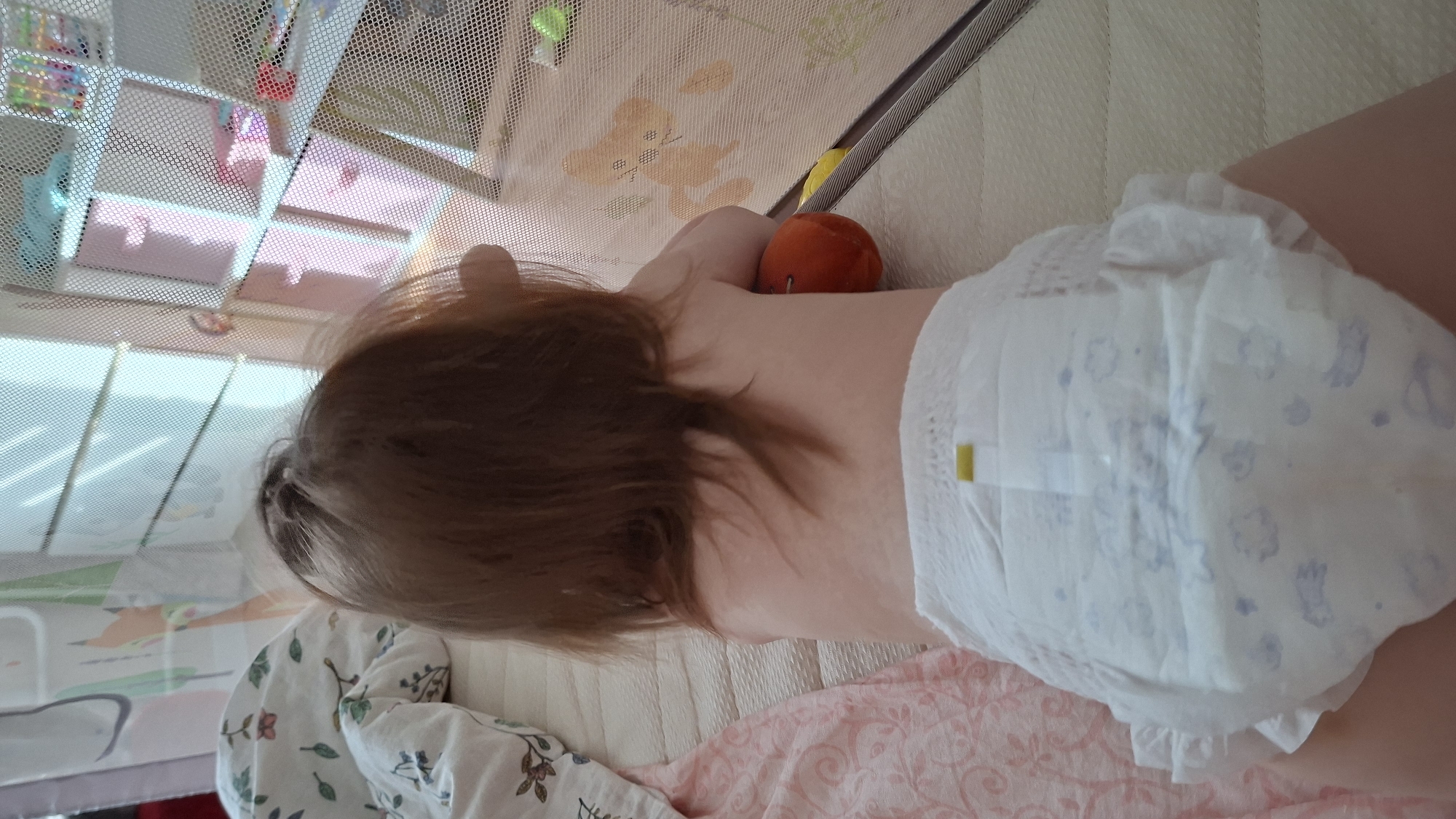 A toddler, seen from the back, lies in a crib in what appears to be a room in an apartment. The crib is covered by a mesh netting. The toddler wears a disposable diaper. An orange fruit rests near the toddler's neck. The room includes a white shelving unit. The location is Chaikovsky, Russia.
