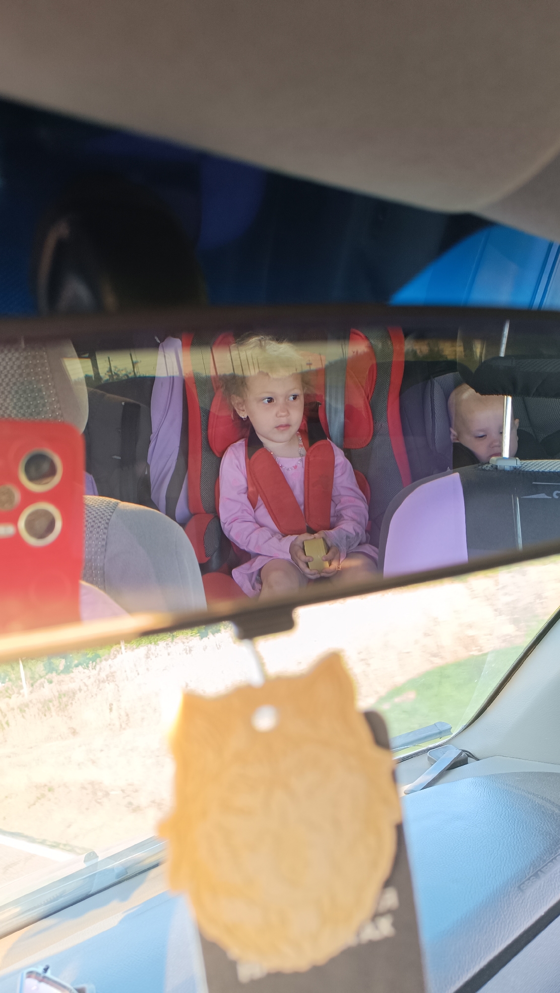 The image shows a rearview mirror reflection of two young children in a car. The child in the foreground, a girl with curly blonde hair, is sitting in a red car seat and wearing a pink long-sleeved dress. She is holding a small yellow object. Behind her, another child, a baby, is visible in a gray car seat. The car's interior is reflected in the mirror, including a red phone case on the left and a furry air freshener hanging in the foreground. The background visible in the reflection appears to be a rural or natural landscape with trees and fields. The metadata indicates the location as Chumna, Russia.