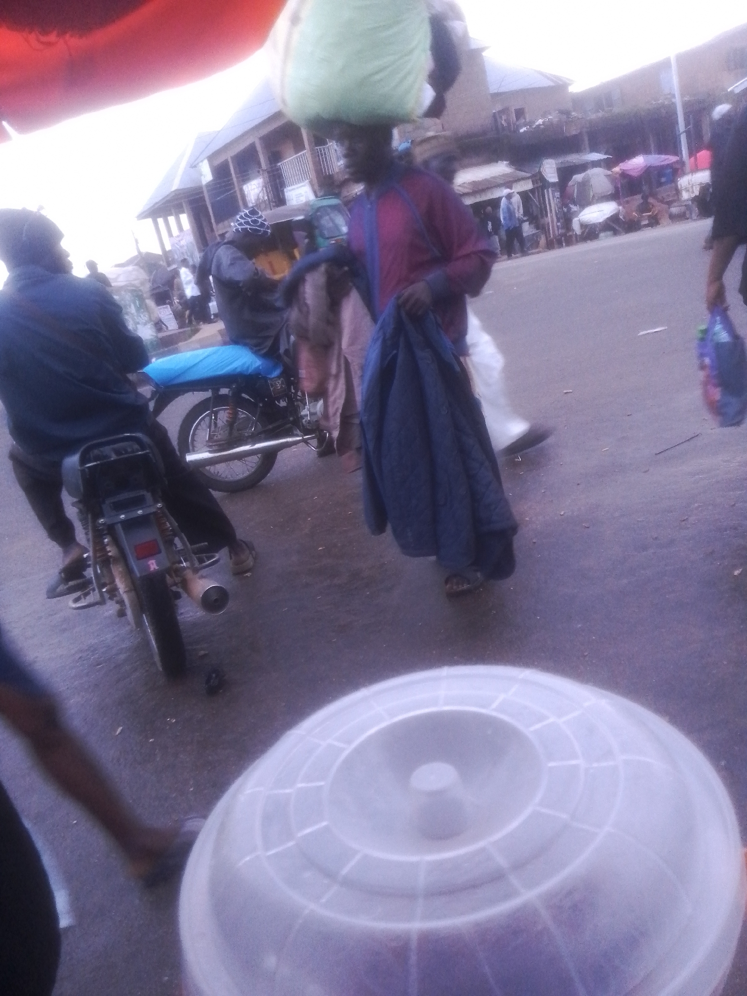 The image depicts a busy street scene, likely in Gwong, Nigeria. A person wearing a dark jacket and black pants is seated on a motorcycle. The motorcycle is black with a blue seat cover and a silver exhaust pipe. Another person, to the left, is wearing a blue jacket and dark pants, and is also on or near a motorcycle. In the background, there are several market stalls with various goods and people. The overall atmosphere suggests a vibrant commercial area.