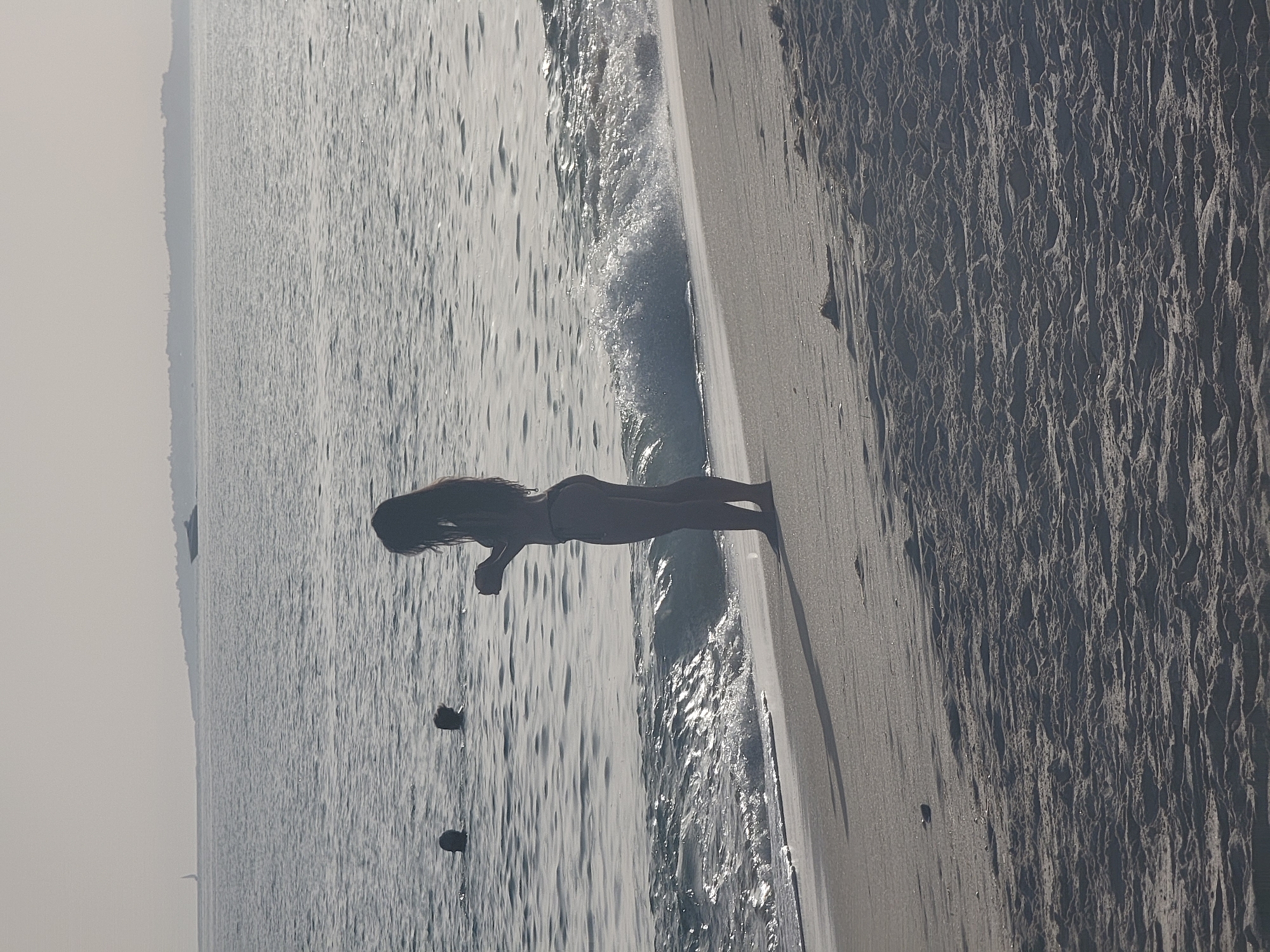 A silhouetted woman in a bikini stands on a sandy beach, facing the ocean. She is holding a small object in her hands. The ocean is calm with gentle waves lapping the shore. In the background, several other people are visible in the water. A distant landmass with a few buildings is visible on the horizon, as well as a boat. The scene appears to be set in Quarteira, Portugal, possibly during daylight hours given the lighting. The sand is dark and textured, showing ripples from the wind or water.