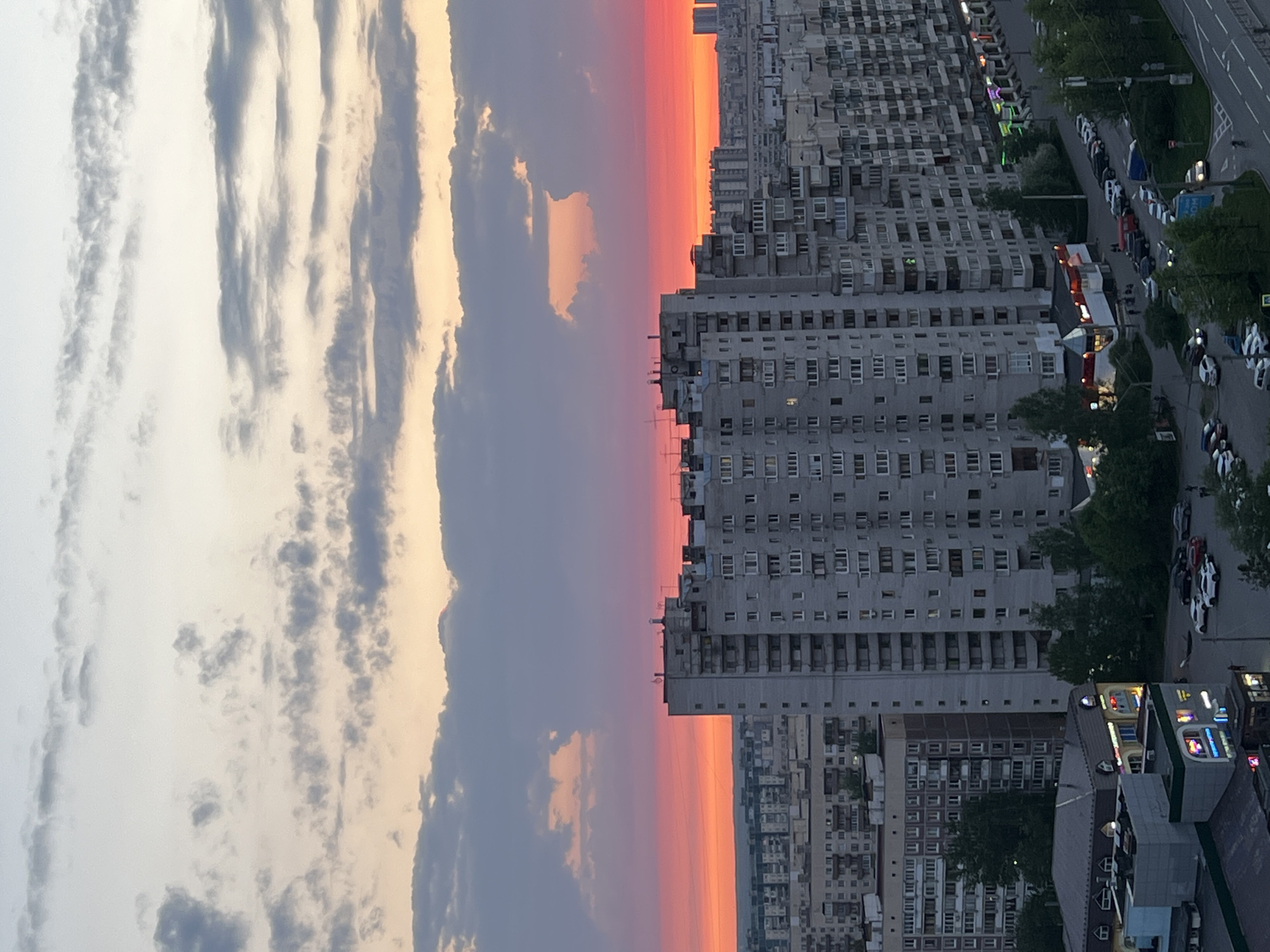The image depicts a high-angle, horizontal view of an urban landscape under a vibrant sunset sky. In the right portion of the image, dense, grey apartment buildings, typical of Soviet-era architecture, dominate the skyline. A street runs alongside the buildings, lined with parked cars and scattered trees. Lower buildings with lit signs indicate commercial activity. The sunset sky transitions from orange near the horizon to purple and grey upwards, with distinct cloud formations in the left portion of the image. The overall impression is of an urban area during twilight. While the buildings evoke the architecture of Saint Petersburg, Russia, confirmation of the location would require additional information or landmarks. There are no people visible in the image.