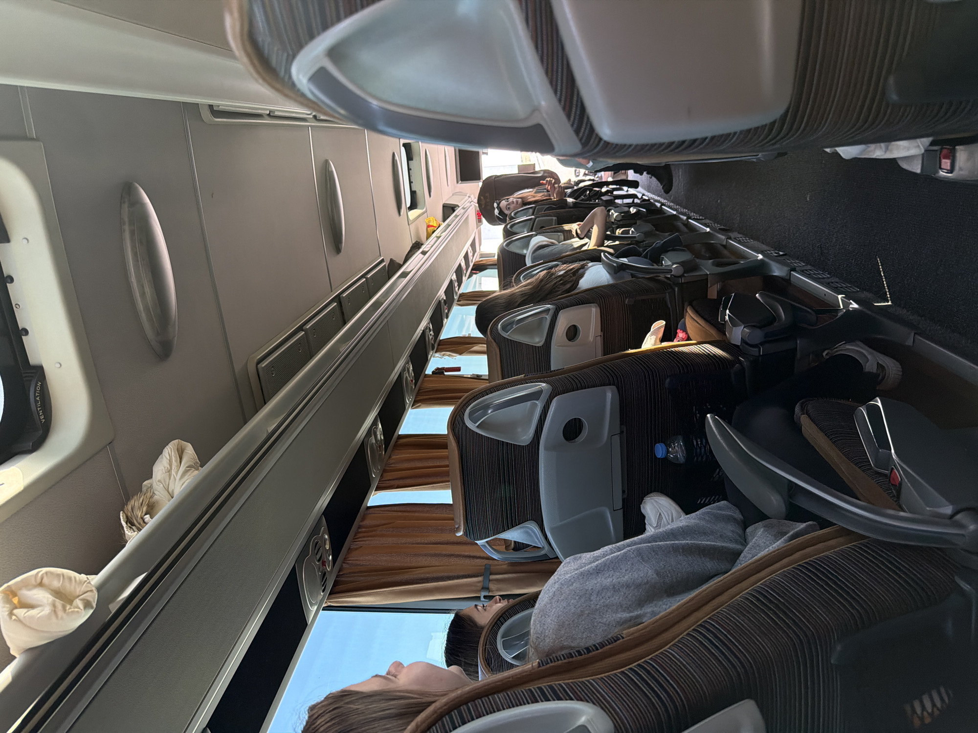 The image depicts the interior of a bus with passengers. The seating arrangement is rows of upholstered chairs. People are seated, some appearing to be resting. The bus windows have curtains, and the landscape visible outside suggests daylight. The interior lighting is soft. The image doesn't give information regarding the location. It is not possible to determine the location in the city of Malko Tranovo, Bulgaria, with the available context.