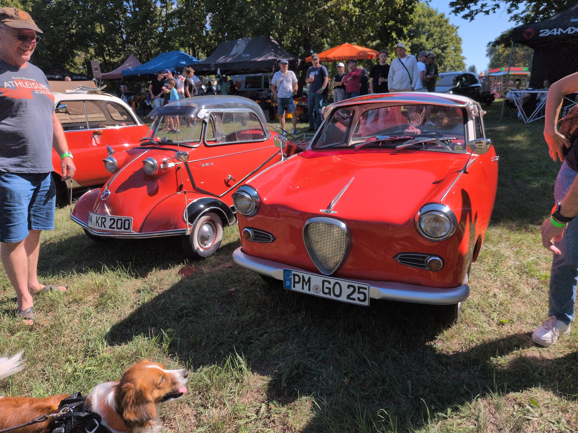 In Hohenferchesar, Germany, at an outdoor event, two red vintage microcars are prominently displayed on a grassy area under a clear sky. A man in a grey t-shirt and blue shorts stands to the left, facing the front of the car closest to him. To the right, a person in blue jeans and pink sneakers gestures towards the car in the foreground. A brown and white dog lies on the grass in the lower left corner of the image, looking towards the right. In the background, tents and several other people are visible, suggesting a public gathering or exhibition. The car in the foreground has a license plate reading "PM GO 25", and the car behind it has a license plate reading "M KR 200". The environment is characterized by green grass and trees in the background.