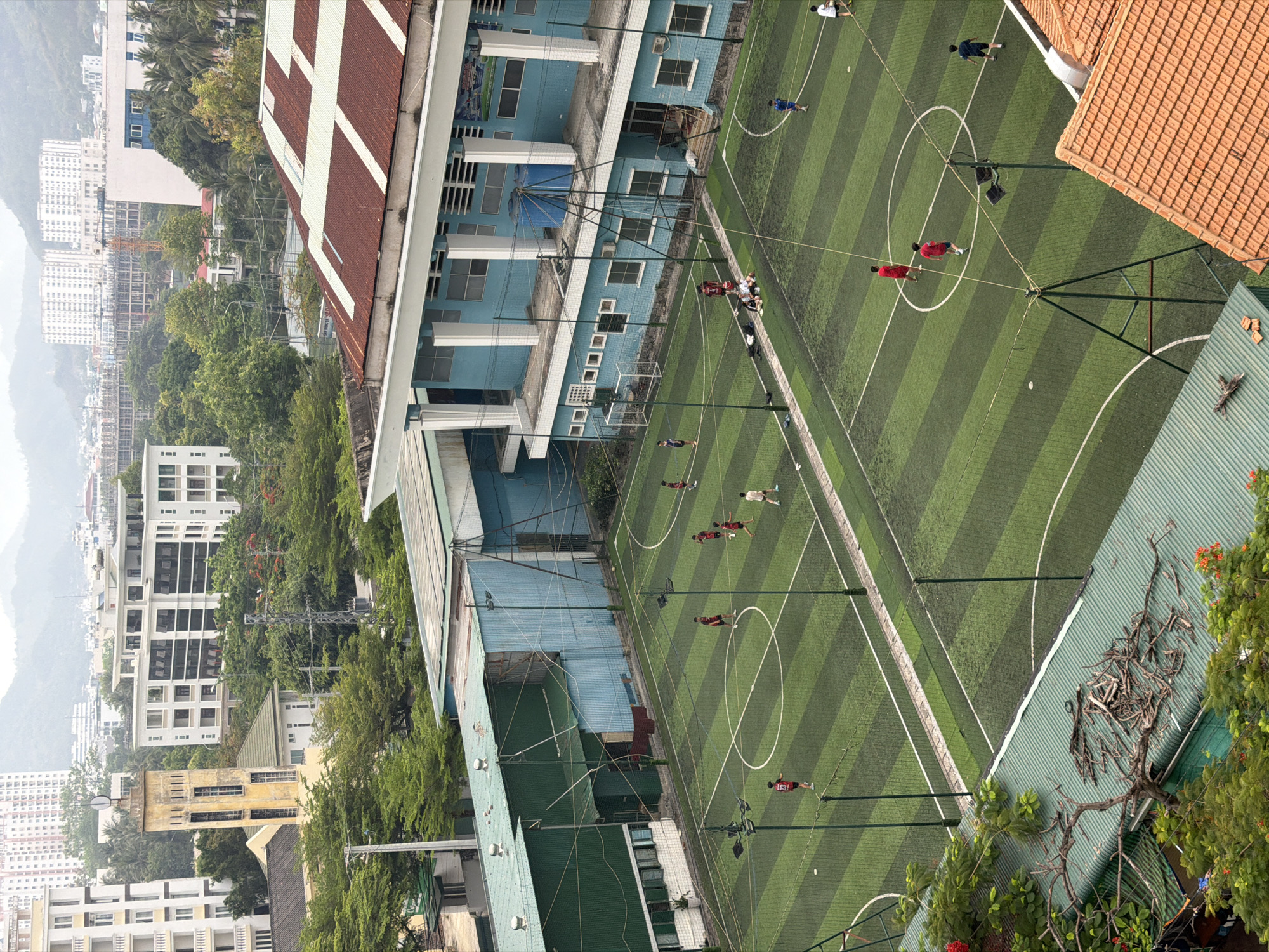 The image shows an elevated view of a soccer field in Bac Nha Trang Ward, Vietnam. A game is in progress with players in red and white uniforms. The field has alternating stripes of dark and light green artificial turf. Tall light posts surround the field. Adjacent to the field is a light blue building with a corrugated metal roof, and a separate building with a red tile roof. Trees and other buildings can be seen in the background. The sky is overcast, and there are mountains visible in the distance.
