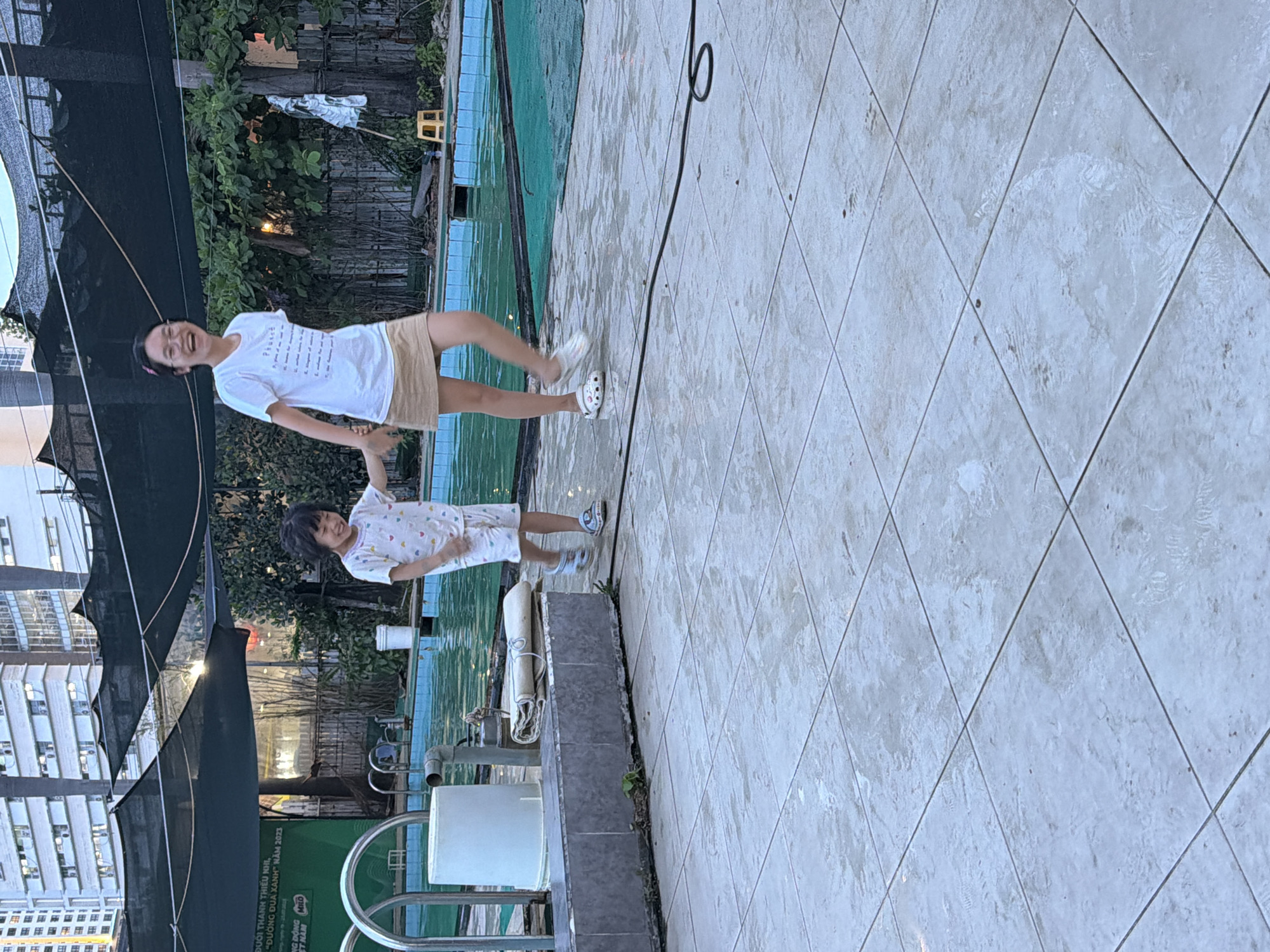 An outdoor scene shows two females, an adult and a child, walking side-by-side on a tiled area next to a swimming pool. The adult, on the right, wears a white t-shirt and a tan skirt, with glasses and a happy expression. She is holding the child's hand. The child, on the left, wears a light-colored pajama set with a pattern and is also smiling. They are near the edge of a swimming pool with blue water. In the background, there are buildings, trees, and netting over the pool area. The tiled ground appears wet, suggesting the area may have recently been rained on or is near the pool. The location is identified as Nha Trang Ward, Vietnam, based on context from a sign visible in the background.