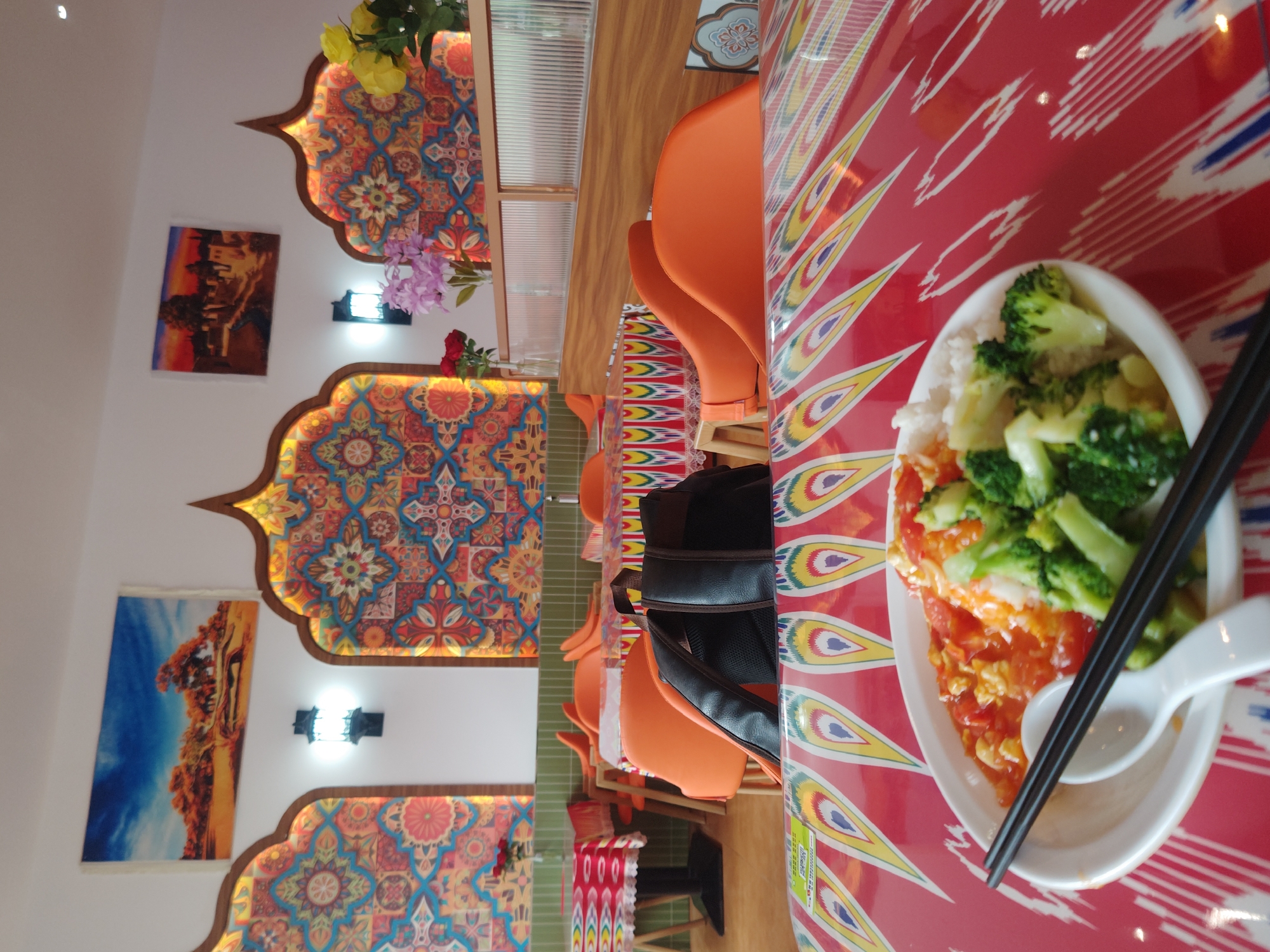 The image displays the vibrant interior of a restaurant, likely located in Chongqing High-tech Zone, China. The scene focuses on a dining table with a distinctive red tablecloth adorned with a pattern of elongated, multi-colored, teardrop-like motifs, reminiscent of Central Asian or Uyghur textiles.
In the foreground, a white oval-shaped bowl holds a meal consisting of white rice, scrambled eggs with tomatoes, and steamed or stir-fried broccoli. A pair of black chopsticks and a white ceramic spoon are placed in the bowl, ready for use. A black backpack is visible resting on one of the orange chairs next to the table.
The background showcases the restaurant's elaborate decor. The walls feature several large, arch-shaped decorative panels with intricate, brightly colored geometric and floral patterns, which appear to be backlit, creating a warm ambiance. Between these ornate panels, rectangular paintings depicting abstract or landscape scenes, characterized by bold colors like blues, oranges, and browns, are mounted. Artificial flowers in yellow, purple, and red are strategically placed atop the wall decorations.
Further into the dining area, rows of simple orange chairs are visible, paired with wooden tables. A section of the wall behind the seating area features green tiling. The lighting throughout the restaurant seems bright and artificial, suggesting an indoor setting independent of natural light. No individuals are visible in the photograph. The decor's style strongly hints at a Central Asian or Silk Road theme, common in ethnic minority restaurants across China. No discernible text is clearly legible in the image.