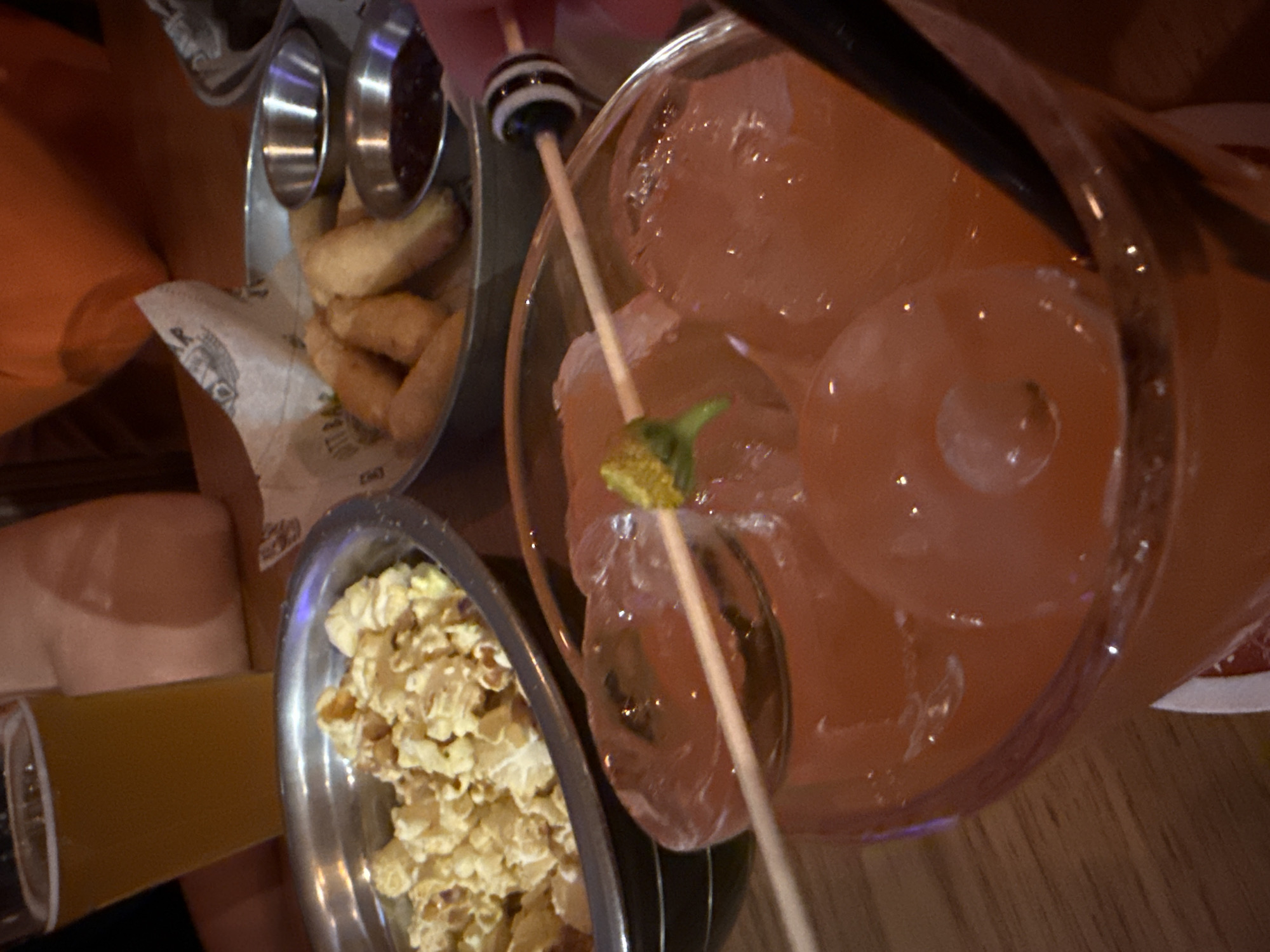 This image captures a close-up, high-angle view of a casual dining or bar scene in Yekaterinburg, Russia, under dim lighting, suggesting an evening setting.
In the foreground and central to the image, a tall glass holds a reddish-orange beverage, likely a cocktail, filled with numerous ice cubes. A black straw is visible within the drink. A wooden skewer, adorned with a black and white striped bead and a small, greenish, textured garnish (possibly a dried fruit or flower bud), is placed in the glass.
To the left, a metal container is filled with golden-brown potato wedges or thick-cut fries, lined with paper featuring faint text, including the word "WINGS." Inside this container are two small, shiny metal dipping cups, one holding a dark sauce. Below this, a metal bowl is filled with popcorn. A partially visible glass containing a yellow liquid, likely beer, is seen on the far left.
A person's hand is visible holding the skewer in the cocktail, and another arm or hand is partially visible on the left side of the frame. The food and drinks are arranged on a wooden table surface. The scene depicts people enjoying snacks and beverages in a relaxed, social atmosphere.