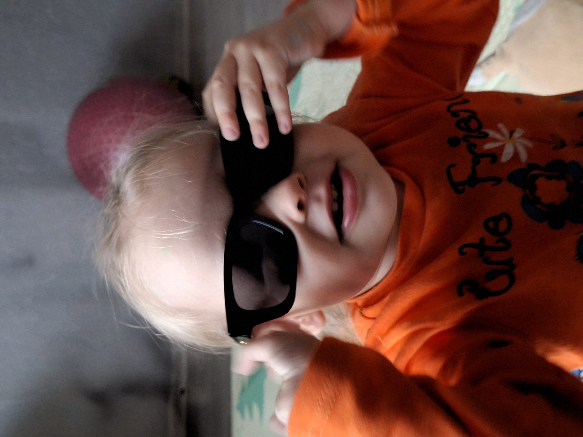A young child with blonde hair is wearing black sunglasses and an orange shirt with the words "Cute Frien" printed on it. The child is holding the sunglasses with their left hand. A pink ball is visible behind the child's head. The setting appears to be indoors. The city and country are unknown.