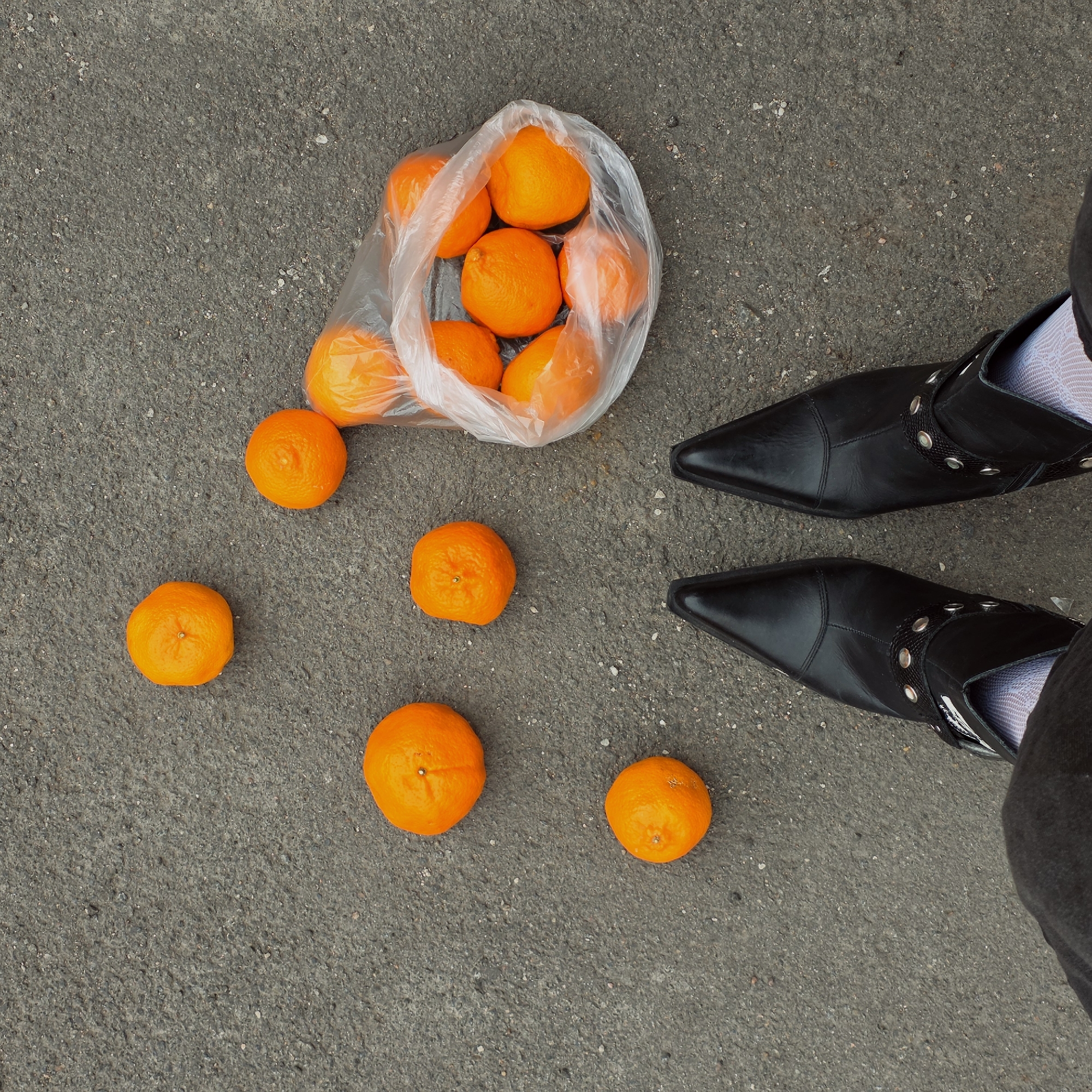 A top-down shot shows a bag of oranges on the ground next to a pair of feet. The oranges are bright orange and are both inside and outside of the plastic bag. The bag sits on an asphalt surface. A pair of black, pointed-toe leather boots with silver rivet details are positioned next to the oranges. A person wearing black pants and white patterned socks is wearing the boots. The location is Saint Petersburg, Russia.