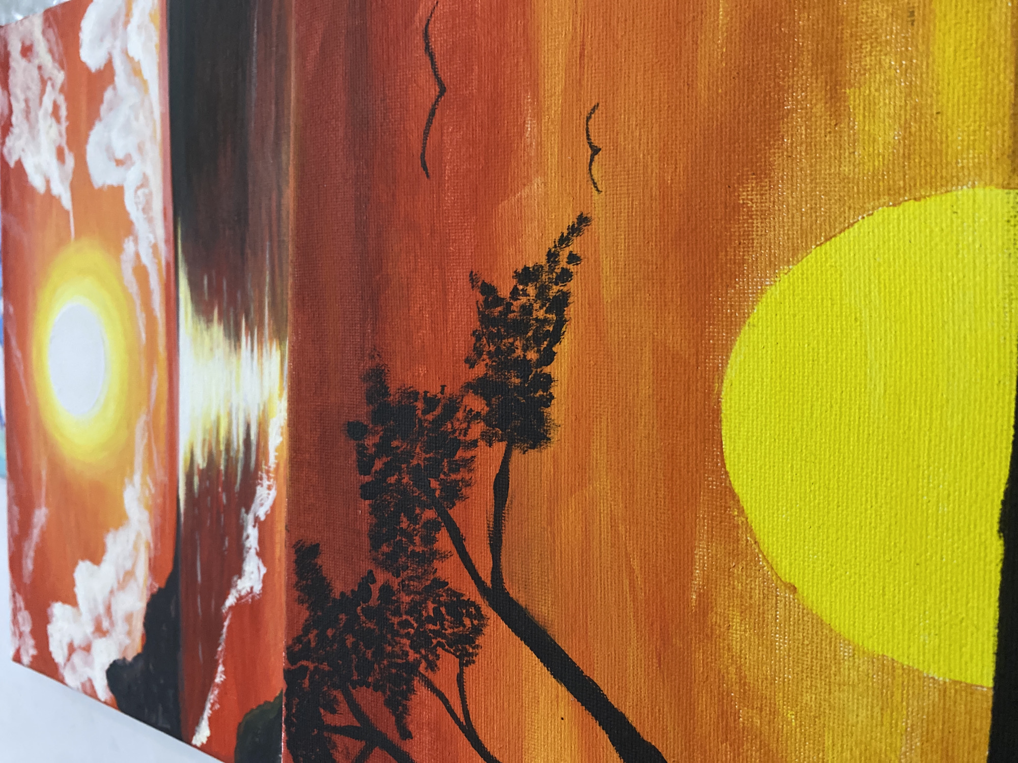 The image displays a series of interconnected landscape paintings, likely executed with acrylic on canvas, presented with a slight angle revealing the thickness of each artwork. These paintings are arranged side-by-side, creating a continuous visual narrative of a natural scene. The dominant color scheme across all pieces is a vibrant gradient of warm tones, including deep reds, various oranges, and bright yellows, strongly suggesting a sunset or sunrise.
On the far left, one painting features a luminous yellow sun with a prominent white halo, surrounded by swirling white clouds against an orange sky. Adjacent to it, another canvas presents a dark, silhouetted foreground, possibly land or mountains, with striking vertical reflections on what appears to be a body of water. The central and right-hand sections of the artwork showcase silhouetted trees with sparse foliage, leaning across the canvases. In the upper part of the central sections, two small, dark bird-like figures are visible against the rich orange and yellow sky. The far-right painting prominently features a large, bright yellow sun, partially visible on the right edge, set against a textured orange and yellow background.
The visible texture of the canvases indicates they are painted artworks. The unified color palette and recurring natural elements—sun, sky, silhouetted landforms, and trees—suggest a cohesive artistic theme focused on the beauty of dawn or dusk. The scenes depict natural landscapes without any discernible human activity. The time of day is clearly either sunrise or sunset, emphasized by the dramatic sky colors. No visible text appears in the image.