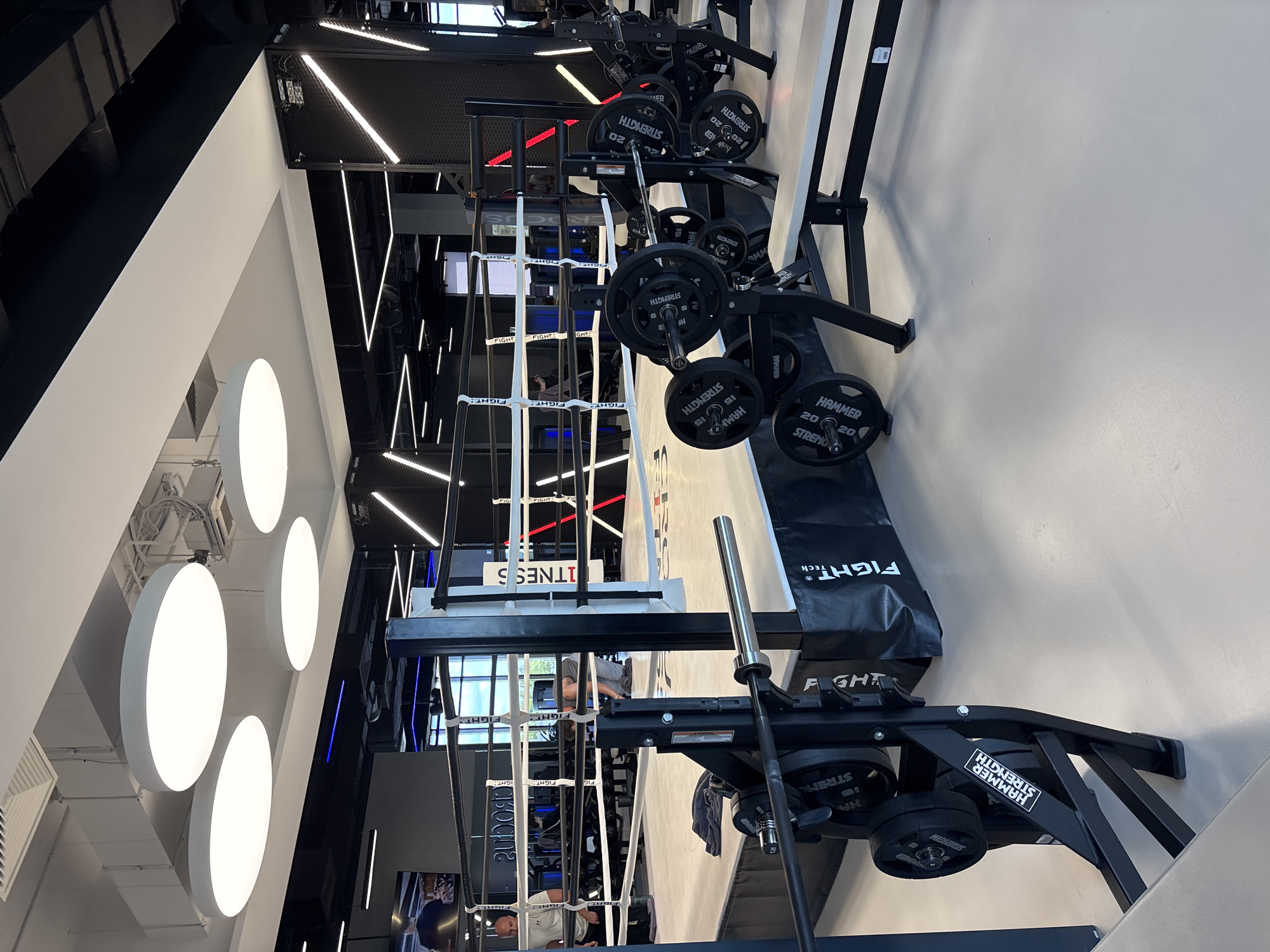 This is an indoor, well-lit gymnasium featuring a boxing ring and weightlifting equipment. The flooring is a light-colored, polished surface. In the foreground, a weight bench with barbells and weight plates is visible. The weight plates are black with white lettering, and some indicate "HAMMER STRENGTH" and weight values like "20". A metal rack with more weight plates is attached to the bench.
The boxing ring is centrally located, with white ropes and black posts. Inside the ring, two individuals are present. One person, wearing a light-colored shirt, stands near the ropes. Another person, wearing a dark tank top, is crouched down on the canvas, appearing to adjust something. The ring is surrounded by a black padded base, with the "FIGHT TECH" logo visible on it.
In the background, additional gym equipment, including racks of weights, can be seen. The ceiling features multiple circular, white fluorescent lights and industrial ductwork. The walls are a mix of black and white panels, with diagonal neon lighting accents. A screen displaying an image of people in a gym setting is mounted on the wall behind the boxing ring. The overall environment suggests a modern and well-equipped fitness facility.