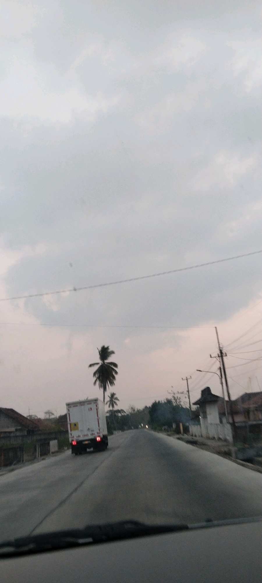 The image depicts a scene from the perspective of a vehicle traveling on a road, likely in Pagar, Indonesia, given the presence of tropical vegetation like coconut palms and the architectural style of the houses visible. The time of day appears to be late afternoon or early evening, indicated by the overcast sky with a hint of pinkish-orange light, suggesting dusk.
In the foreground, the dashboard and windshield wipers of the vehicle are visible, framing the view ahead. A white truck with red taillights is traveling in the same direction on the road, positioned to the left of the frame. The truck has some markings on its rear, including what appears to be a yellow sticker with black text.
The road itself is paved and appears to be a two-lane highway. On either side of the road, there are signs of habitation, with houses visible on the left and right. The houses on the left have tiled roofs and are partially obscured by vegetation. On the right, there are more structures, including what looks like a utility pole with numerous wires stretching across the sky.
The environment is characterized by lush greenery, with prominent coconut palm trees dotting the landscape. The sky is filled with a thick layer of clouds, giving the scene a diffused light. A few smaller trees and bushes are also visible along the roadside. The overall impression is one of a rural or semi-urban setting in a tropical region. There are no people clearly visible in the image, and the primary subjects are the road, the truck, and the surrounding environment. The image conveys a sense of movement, as if the viewer is on a journey.