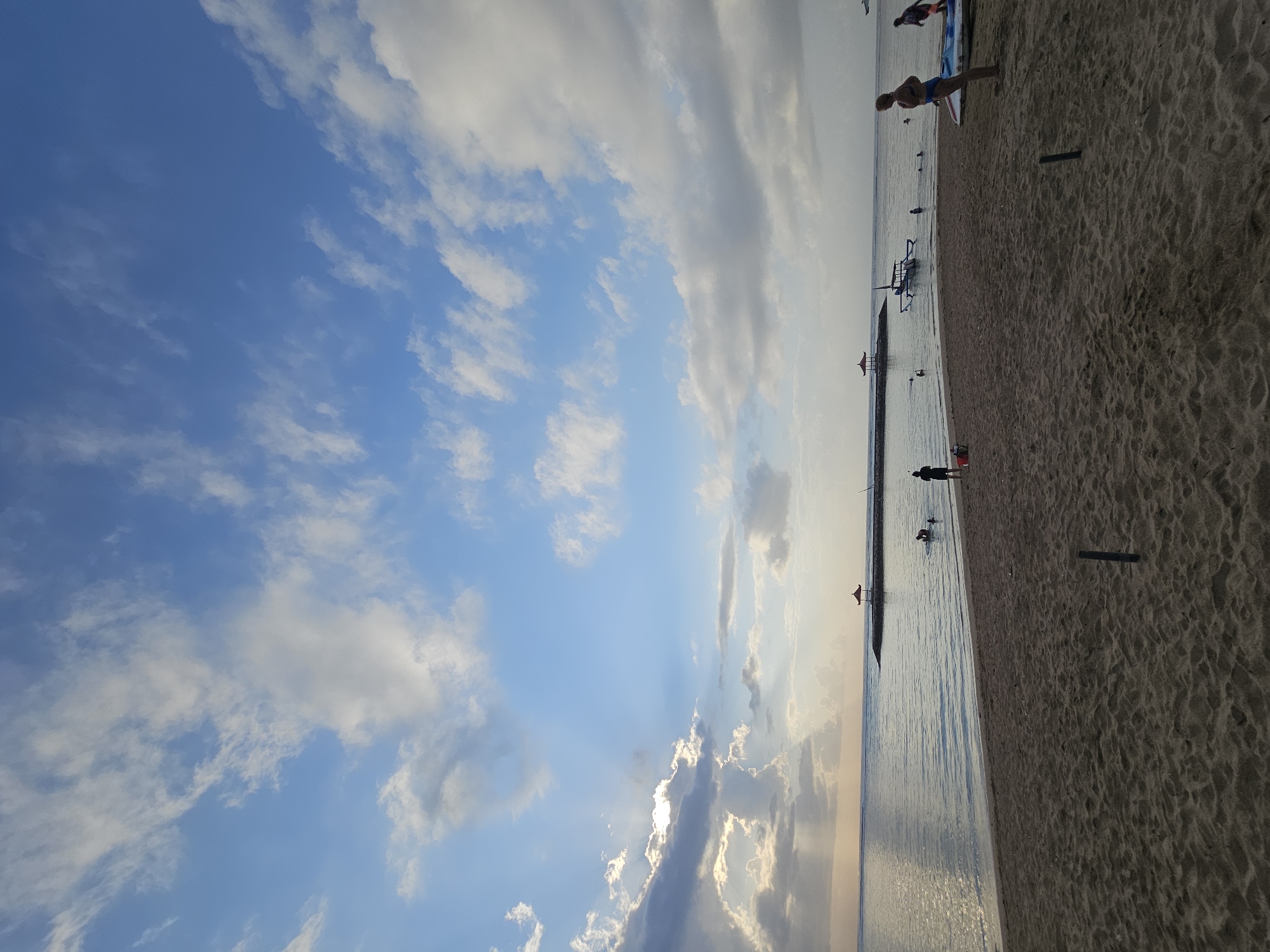 A wide-angle, vertical, eye-level shot shows a sandy beach in Denpasar, Indonesia, with the ocean and sky dominating the scene. The sand in the foreground is a light brown color, with some small dark poles visible. The ocean is calm and reflects the sky, with a breakwater made of stones extending into the water. On the breakwater, there are two small, red-roofed gazebos. Several people are in the water, some swimming and some standing. A small boat with a blue hull is moored near the breakwater. On the far right of the beach, a woman in a blue bikini stands next to a surfboard and a kayak. In the distance, the sun is setting, casting a warm glow on the clouds. The sky is a brilliant blue, with scattered white clouds.
