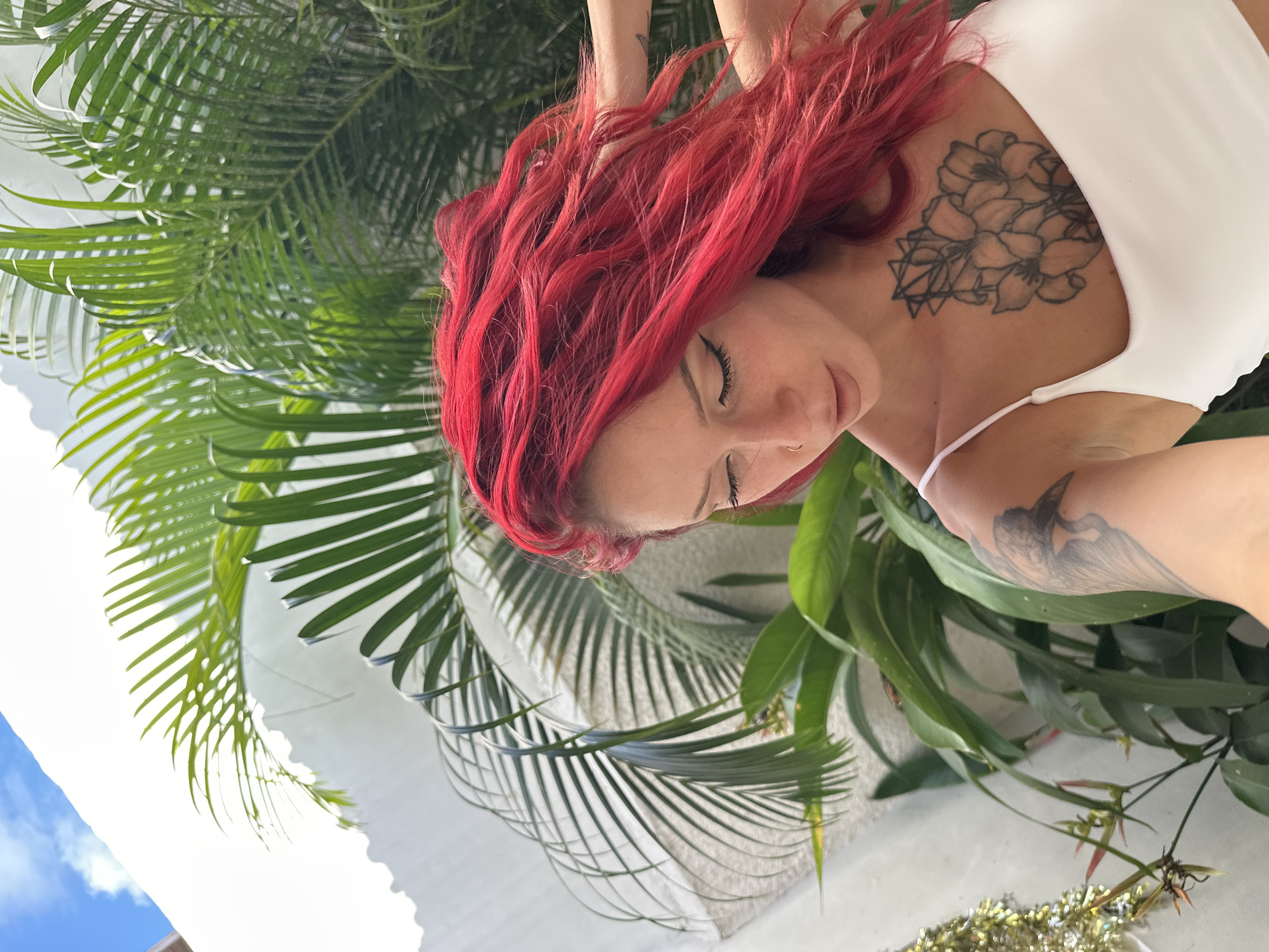 A woman with vibrant red hair, a white top, and tattoos on her upper chest and arm is the focal point of this angled shot. The woman's eyes are closed. She is positioned amidst lush green foliage, including palm leaves and other tropical plants. The background includes a glimpse of a white building under a bright sky with soft clouds. This image seems to be taken in an outdoor setting, possibly a garden or near a building exterior, North Kuta, Indonesia.