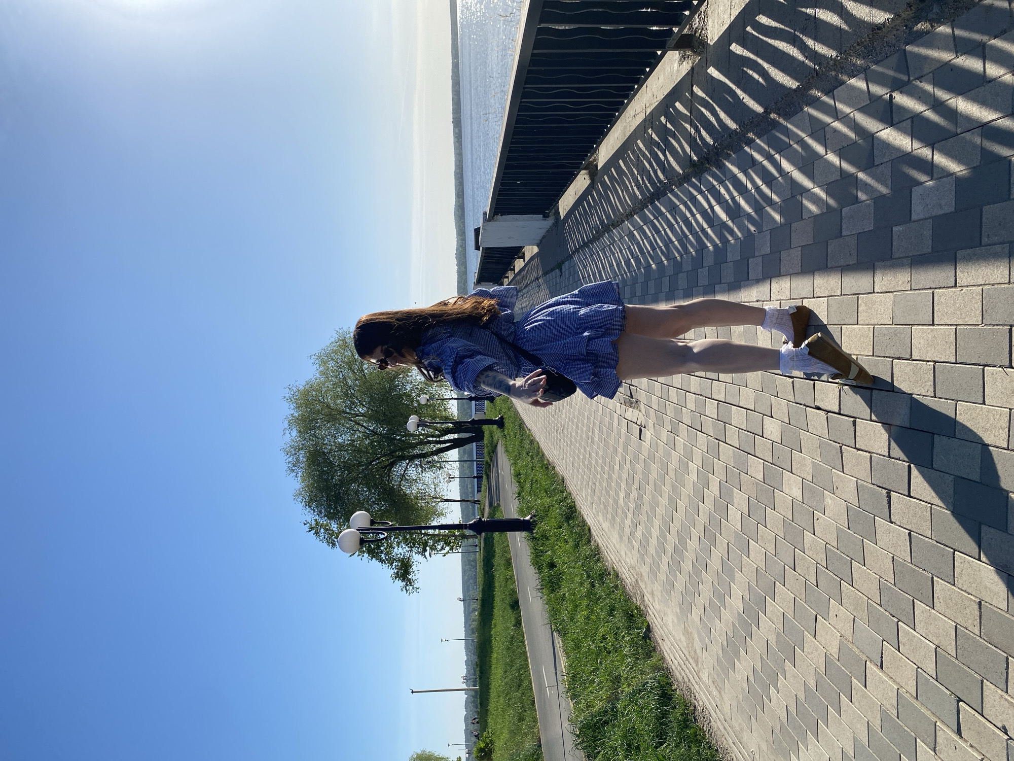 A woman with long brown hair is standing on a paved walkway in Votkinsk, Russia. She is wearing a short blue and white gingham dress, a denim jacket, white socks, and brown shoes. The walkway is made of gray paving stones and runs along a wrought-iron fence. Beyond the fence is a body of water. To the left of the woman, along the walkway, there are street lamps and greenery. The sky is blue.