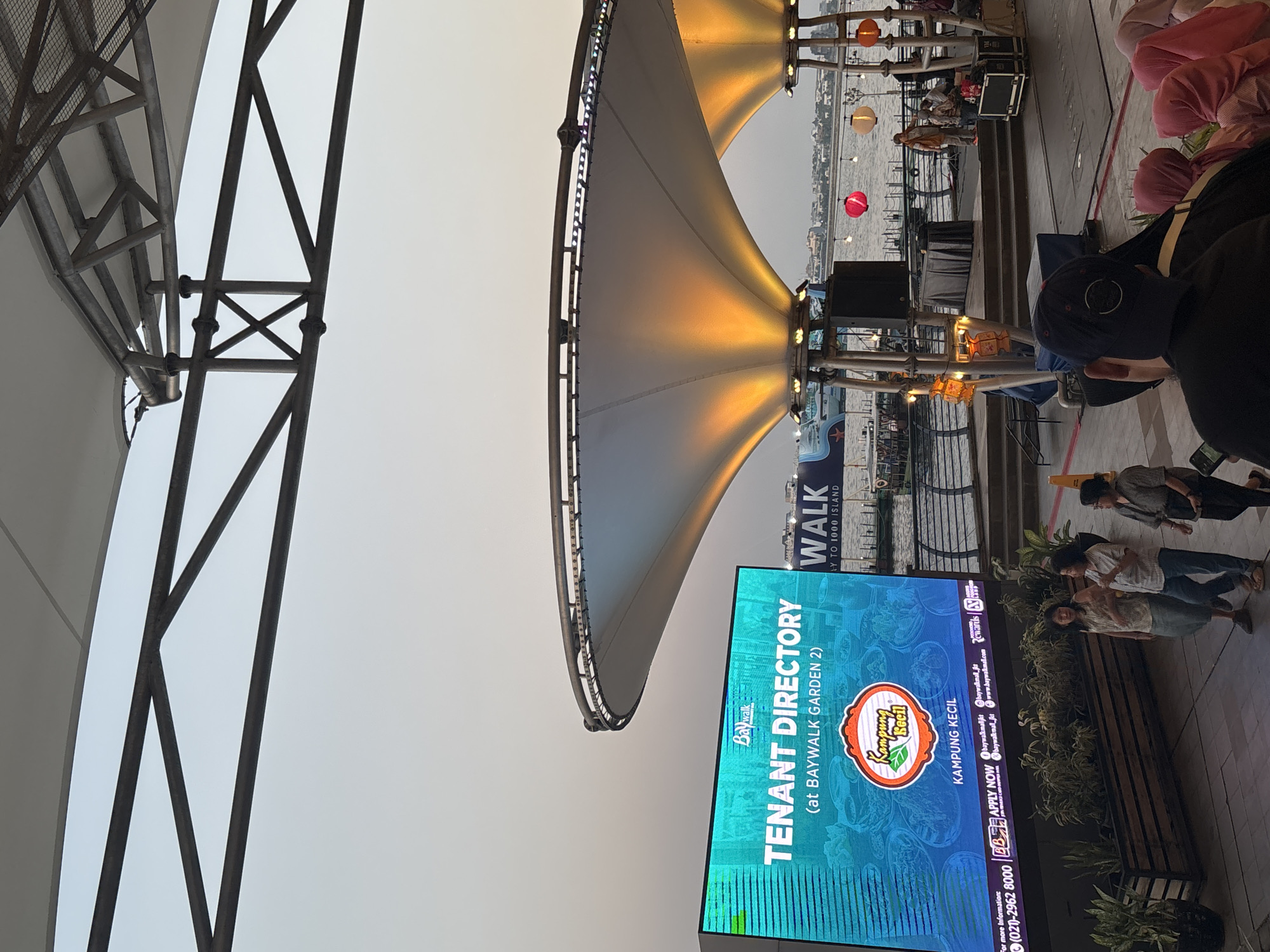 The image depicts an outdoor, waterfront scene at dusk or early evening, identified as Baywalk Mall in North Jakarta, Indonesia, with a sign indicating "BAYWALK GARDEN 2" and "TO 1000 ISLAND" in the distance.
A prominent, large LED screen is positioned on the left, displaying a "TENANT DIRECTORY (at BAYWALK GARDEN 2)" with the Baywalk logo. Below this, a logo for "Kampung Kecil" is featured, followed by contact information including a phone number "(021)-2962 8000" and social media/website addresses. The screen's background shows blurred images of food. In front of the screen, several dark planters contain green and variegated foliage.
Above the mid-ground, two large white tensile canopies, supported by a metal truss framework, are internally illuminated with an orange light. A section of the metal truss structure is visible across the top of the frame. Beyond the canopies, a body of water is visible, with a dark blue boat featuring a white star logo partially visible on the left. A stage-like area, elevated above the water and enclosed by metal railings, features a black draped table, a large black speaker box, and flight cases. Several decorative lanterns (red, beige, orange) are suspended on a line above the water.
In the immediate foreground, a person wearing a dark baseball cap is seen from behind, facing away from the camera. To their right, several individuals, mostly women wearing pink or light-colored headscarves, are seated and facing away. In the mid-ground, to the left of the LED screen, three women stand conversing. The leftmost woman has long dark hair, wearing a light patterned top and a dark skirt. The central woman has short dark hair, wearing a striped shirt and jeans. The rightmost woman wears glasses, a dark top, dark trousers, and carries a dark bag. The sky transitions from a light blue at the zenith to a pale orange near the horizon.
