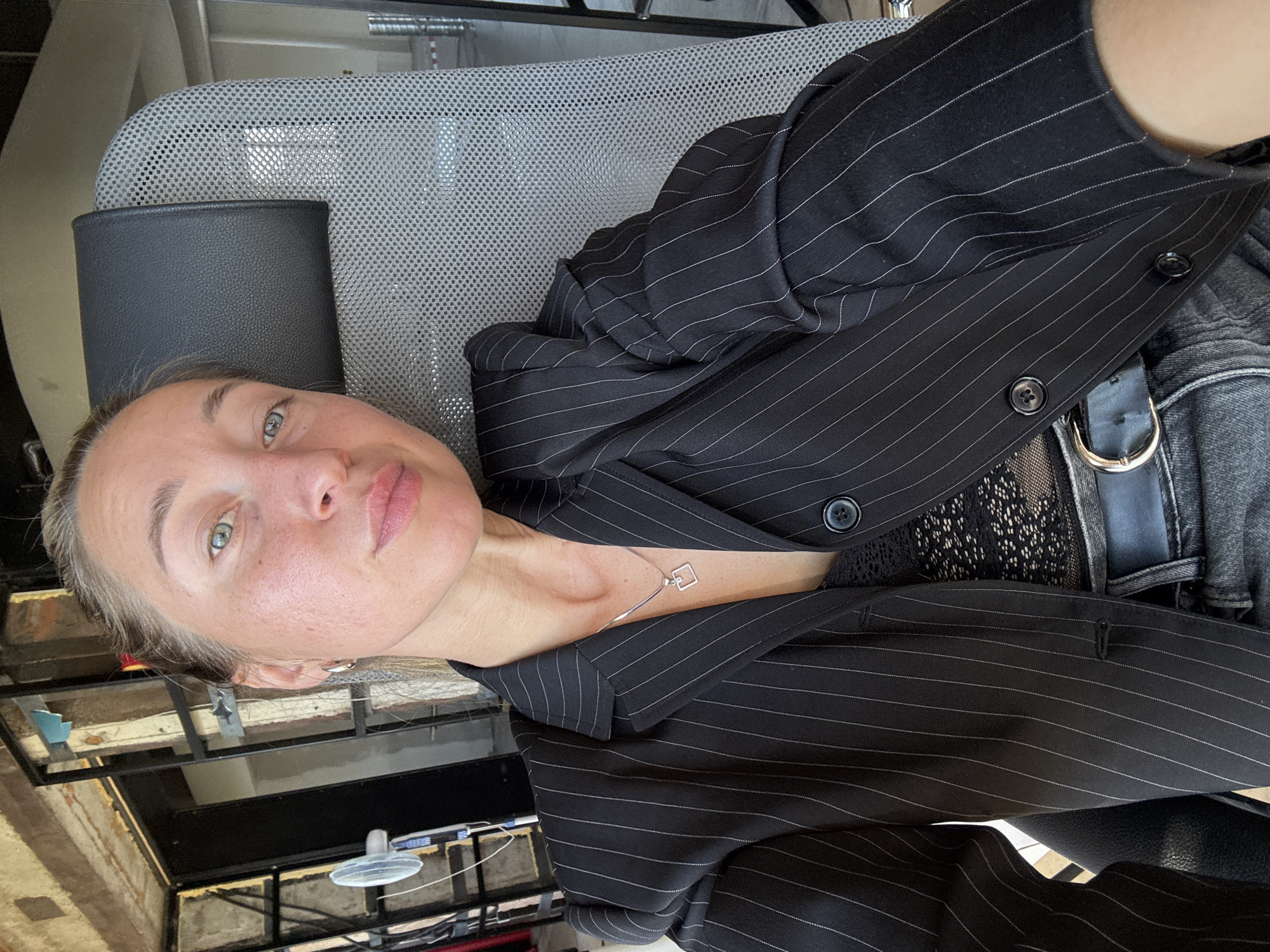 A woman is seated in an office chair, looking at the camera. She has blonde hair pulled back and is wearing a black pinstripe blazer over a black lace top and denim jeans. A silver pendant necklace is visible around her neck. The background appears to be an industrial or unfinished space with exposed brick and metal structures. There is no indication of the specific location within Chaikovsky, Russia.