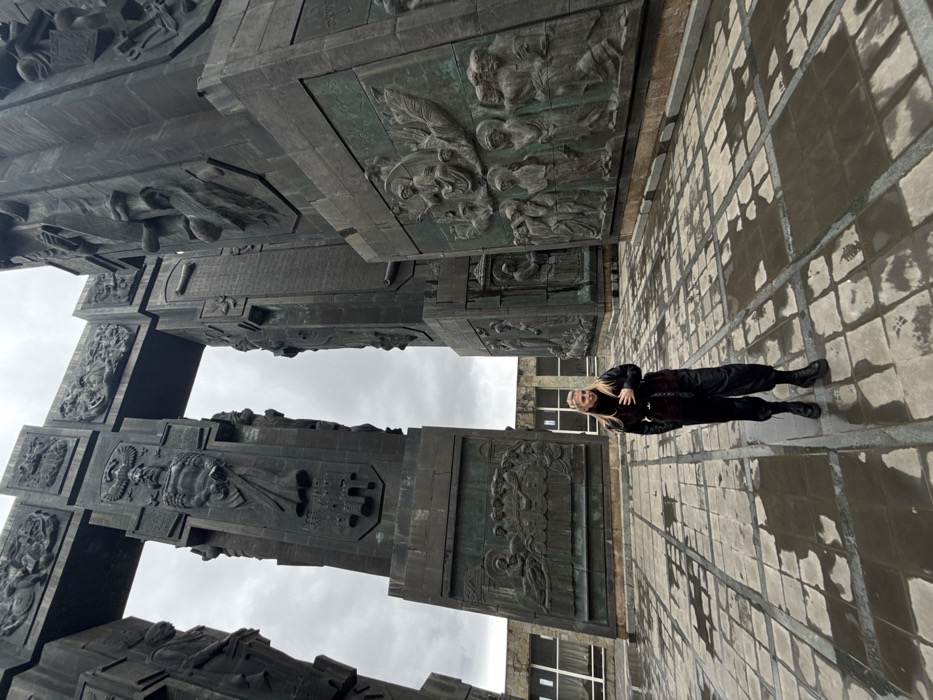 A blonde female stands in the foreground, centered slightly left, amidst the massive bronze columns of the Chronicle of Georgia monument, located on a hilltop overlooking the Tbilisi Sea in Tbilisi, Georgia. She is dressed in a dark, possibly black, ensemble consisting of a long-sleeved top, a dark vest with a textured collar, loose-fitting dark pants, and sturdy black boots. Her hands are positioned across her midsection, and she faces slightly to her left (viewer's right) with a mild expression.
The ground is paved with large square tiles, exhibiting wet patches and reflections from puddles, suggesting recent precipitation. The monument itself comprises towering, dark, patinated bronze columns adorned with numerous intricate bas-relief sculptures depicting historical events, religious scenes (including a representation of "The Last Supper" visible on one panel), and significant Georgian figures. These columns rise high into an overcast, grey sky. In the distant background, behind the monument's structures, a modern building with multiple rectangular windows is partially visible.