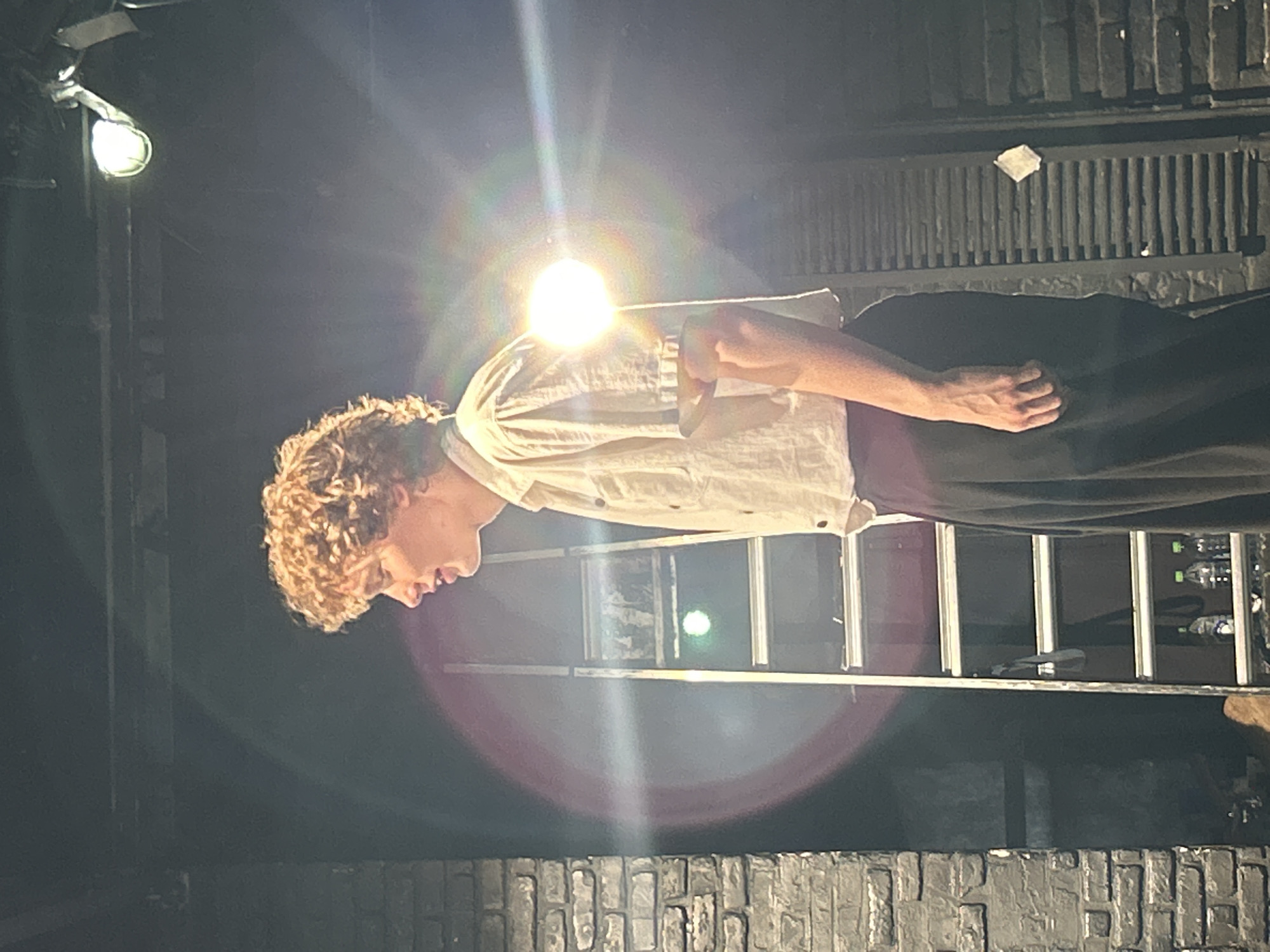The image shows a young person with curly, blonde hair, wearing a light-colored short-sleeved shirt and dark pants. They are facing left, with their head bowed and eyes closed, appearing contemplative or in thought. The person is lit by a bright spotlight from above, which creates lens flares and halos of light.
Behind the person, a metal ladder is visible, suggesting a stage or performance setting. There are also some water bottles and other objects on or near the ladder, possibly props. The background is dark, with hints of brickwork at the bottom of the frame. The lighting and the person's posture create a dramatic and possibly emotional atmosphere. The overall impression is that of a moment captured during a performance or rehearsal, emphasizing the individual's introspective state. The time of day is not discernible, but the strong artificial lighting suggests an indoor or nighttime event. There is no visible text in the image.