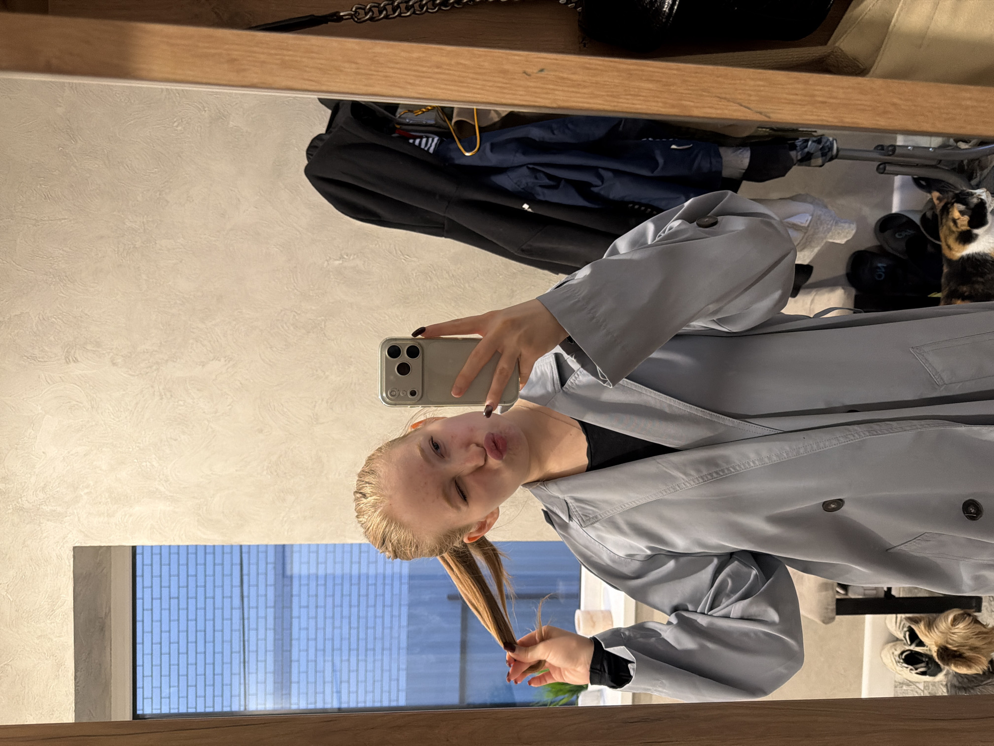 A young woman is taking a selfie in a mirror. She is wearing a light gray trench coat and has her blonde hair pulled back in a ponytail. She is pouting and winking at the camera. The background of the image includes a window with a view of a brick building, a closet with clothes and shoes, and a cat. The location is likely an indoor space in Rossiyskiy, Russia.