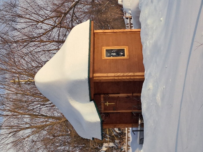 The image displays a small, single-story wooden building, possibly a chapel or shrine, situated in a snow-covered landscape in Полянки, Russia. The structure features light brown wooden plank walls and a steeply pitched, rounded roof heavily blanketed with thick, pristine white snow. A golden Orthodox cross, identifiable by its slanted lower bar, is positioned at the apex of the snow-covered roof. The visible facade includes a wooden door on the left, adorned with a smaller golden Orthodox cross and dark, ornamental metalwork above it. To the right of the door, a single rectangular window with a light-colored wooden frame and a dark, reflective pane is present. The ground surrounding the building is entirely covered in deep, undisturbed white snow, with subtle shadows indicating direct sunlight. In the background, numerous bare deciduous trees rise against a clear, light blue sky, confirming a winter setting. To the left of the building, a dark, low-lying object is partially obscured by snow, and to the right, a wooden fence, also partially covered by snow, extends into the mid-ground. No individuals are present in the scene.