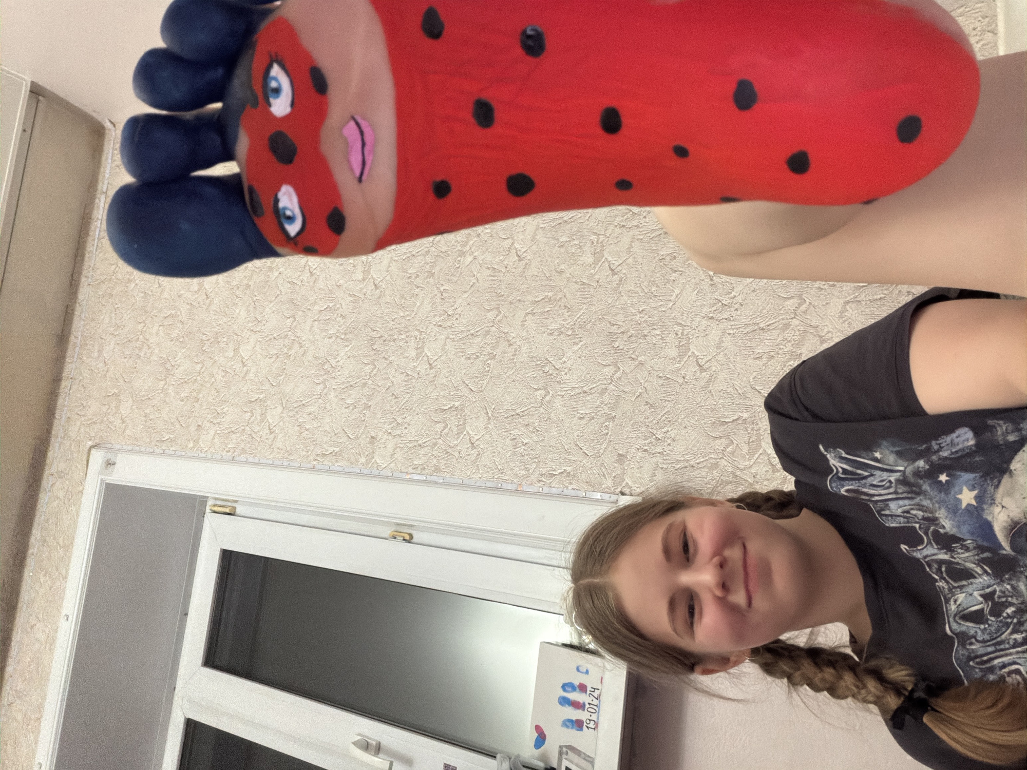 A fair-skinned young woman with blonde hair in braids is holding up her painted foot. The foot is painted to resemble the character Ladybug from Miraculous: Tales of Ladybug & Cat Noir. The toes are painted dark blue, and the rest of the foot is red with black spots and a face. The woman is wearing a dark t-shirt. Behind her is a textured beige wall and a white-framed window. A box with colorful fingerprints and the date "19-01-24" is on the window sill. The location is potentially Perm, Russia.