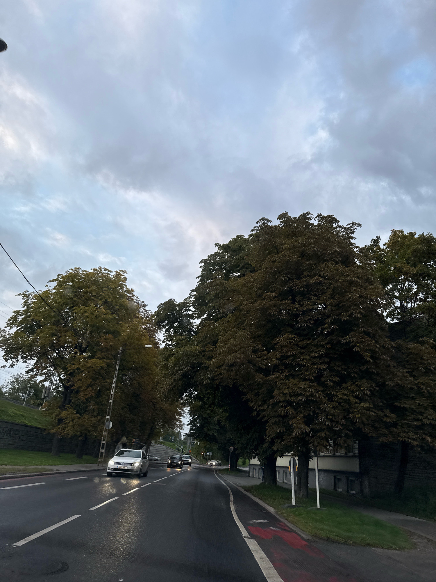 The image depicts a cloudy, overcast sky above a street in Tallinn, Estonia. The road is wet, suggesting recent rain, and traffic is light, with a few cars visible. Lush green trees line the side of the road, their leaves showing hints of autumn colors. The overall atmosphere is subdued and somewhat somber, fitting for a cloudy day in the city.