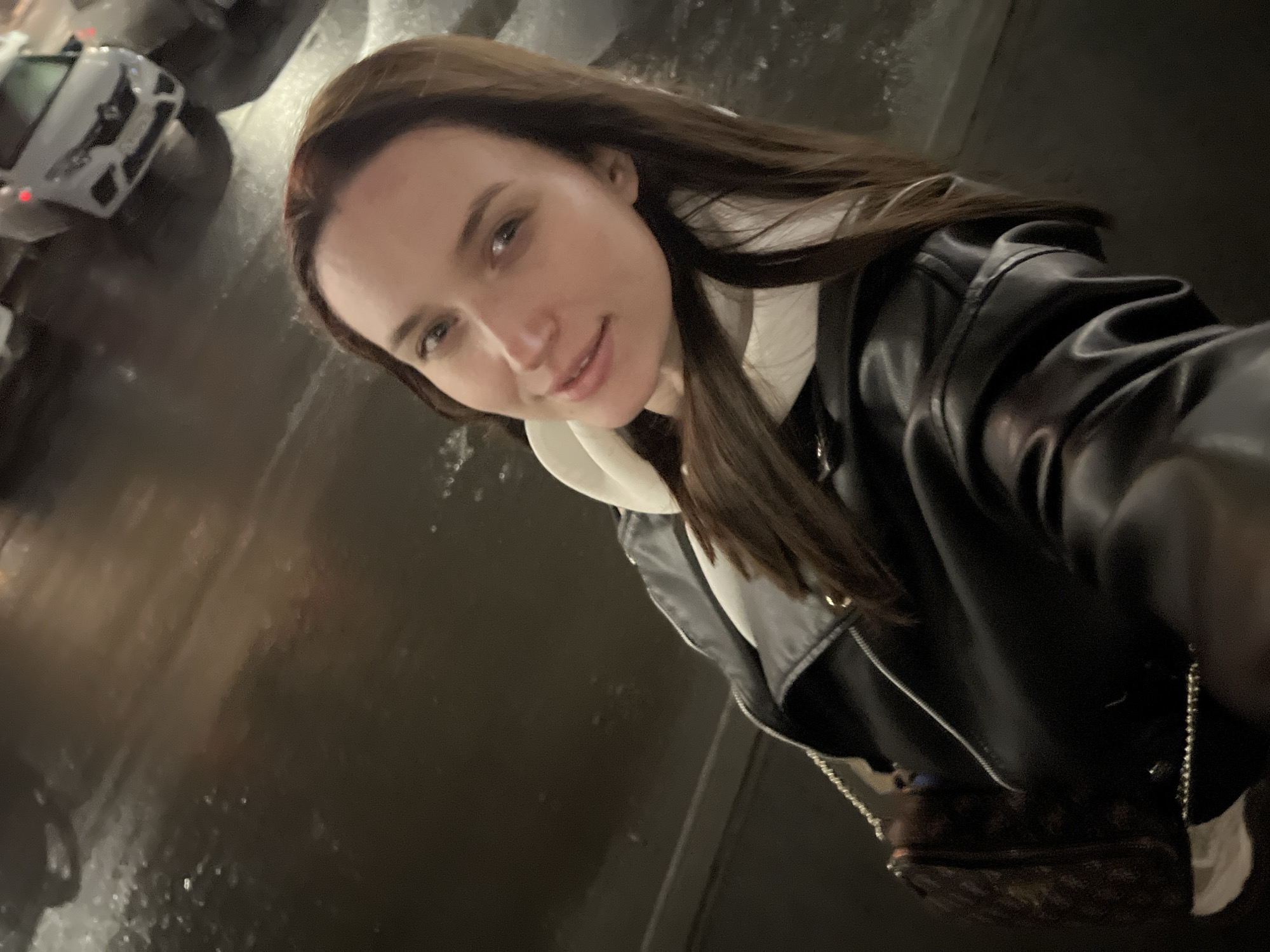 A woman poses for a selfie outdoors at night. Rain appears to be present, as the ground appears wet and reflective. The woman is wearing a black leather jacket over a white hooded sweatshirt. She also has a small brown purse with a chain strap. A vehicle is visible in the background on the left side of the frame. The location is consistent with an urban area, potentially Tyumen, Russia, though no specific landmarks confirm the location.