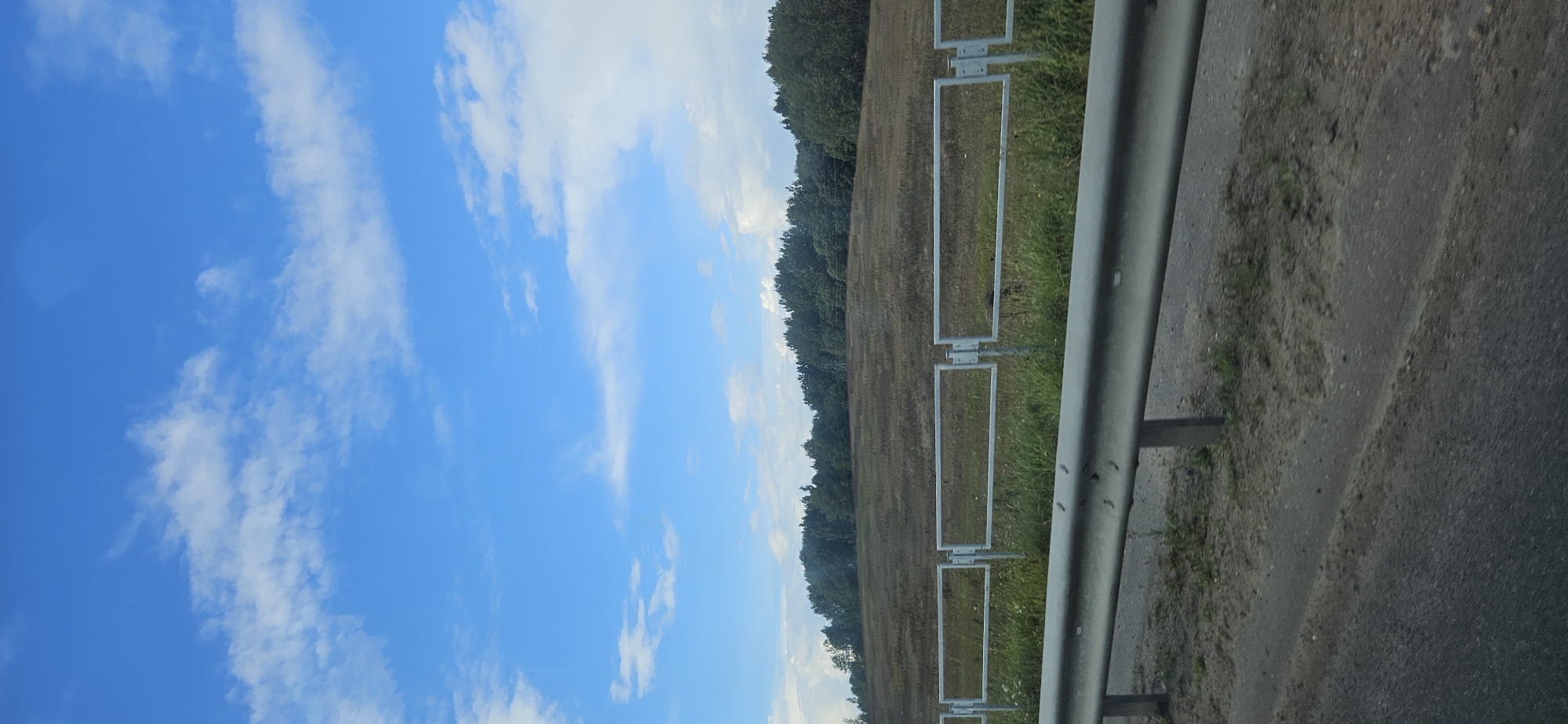 This is a vertical, eye-level shot of a rural landscape under a blue sky with scattered white clouds. In the foreground, a grey metal guardrail runs horizontally across the lower portion of the frame, separating the road from a field. The field itself appears dry and brown, with scattered patches of green grass near the guardrail. Beyond the field, a dense line of dark green trees forms a horizon. The overall scene suggests a tranquil, possibly summer, day in a rural setting. The location of Варонаўскі сельскі Савет, Belarus, is noted as the context for this image.