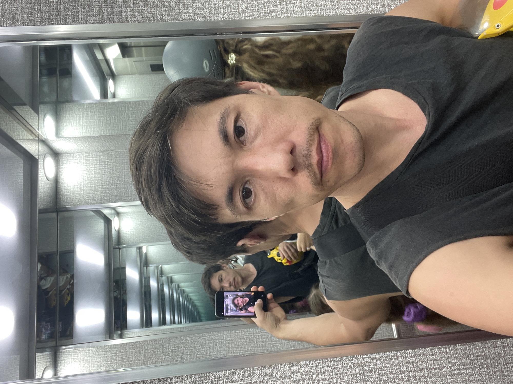 A man is taking a selfie in front of a mirror that reflects an infinite number of images of an elevator interior with interior lights. He has short, dark hair, fair skin, and a light mustache. He is wearing a dark gray t-shirt. He is holding up a smartphone which is displaying an image that is a selfie of a woman. In the reflection in the mirror, a second man and a child can be seen, as well as a yellow object. The reflective surface is bordered by a gray textured wall. The elevator interior is lighted with fluorescent lights. This photo was taken in Bac Nha Trang Ward, Vietnam.
