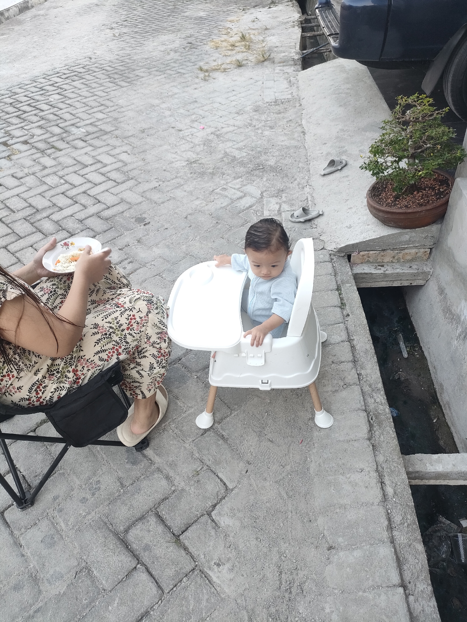 In a paved outdoor area in Unknown, Indonesia, a baby sits in a white high chair. The baby is wearing a light blue onesie and is looking down. A woman in a floral-patterned dress and light-colored slip-on shoes sits on a black folding chair to the left, holding a plate of food. The ground is paved with gray interlocking bricks, and a concrete pathway runs alongside a dark, open drain. A potted bonsai tree sits on the concrete pathway near a dark-colored vehicle. Several grey flip-flops are scattered on the ground near the bonsai tree.