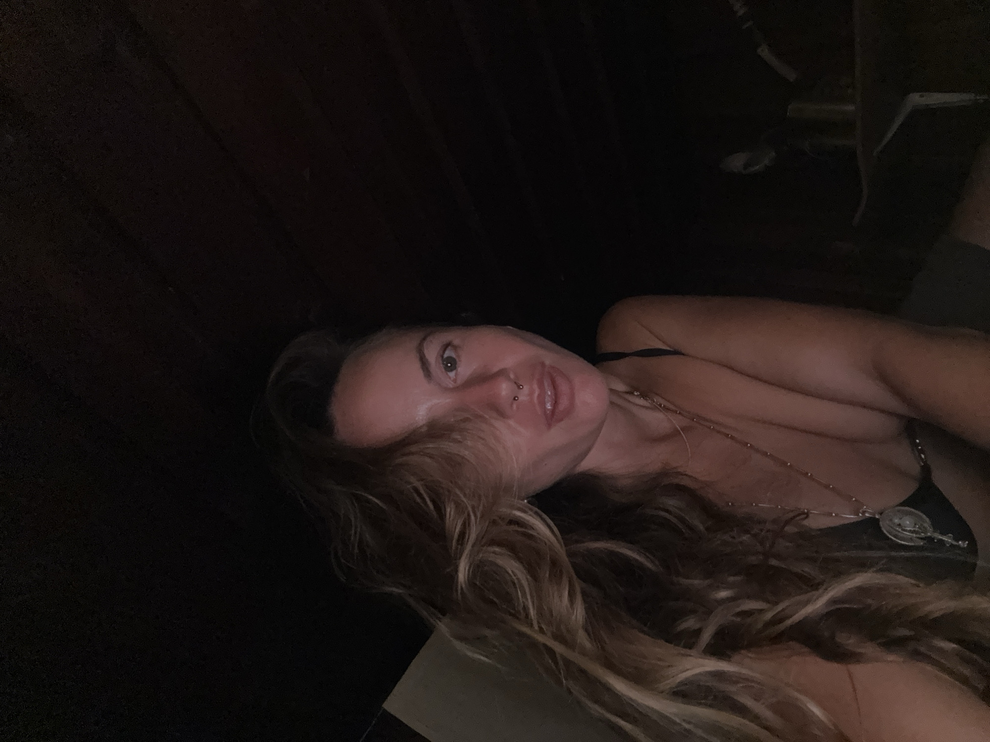 A close-up, low-angle shot captures a woman from the chest up, positioned in the lower left quadrant of the frame. She has long, wavy blonde hair that drapes across her face and shoulder, partially obscuring her left eye. Her head is tilted slightly towards the camera, and her lips are slightly parted. She is wearing a black top and a necklace with a pendant. A nose ring is visible. The background is dark and indistinct, with wooden beams or planks visible in the upper portion of the image, suggesting an indoor or covered outdoor space. The lighting is dim, with a warm tone highlighting the woman's face and hair.