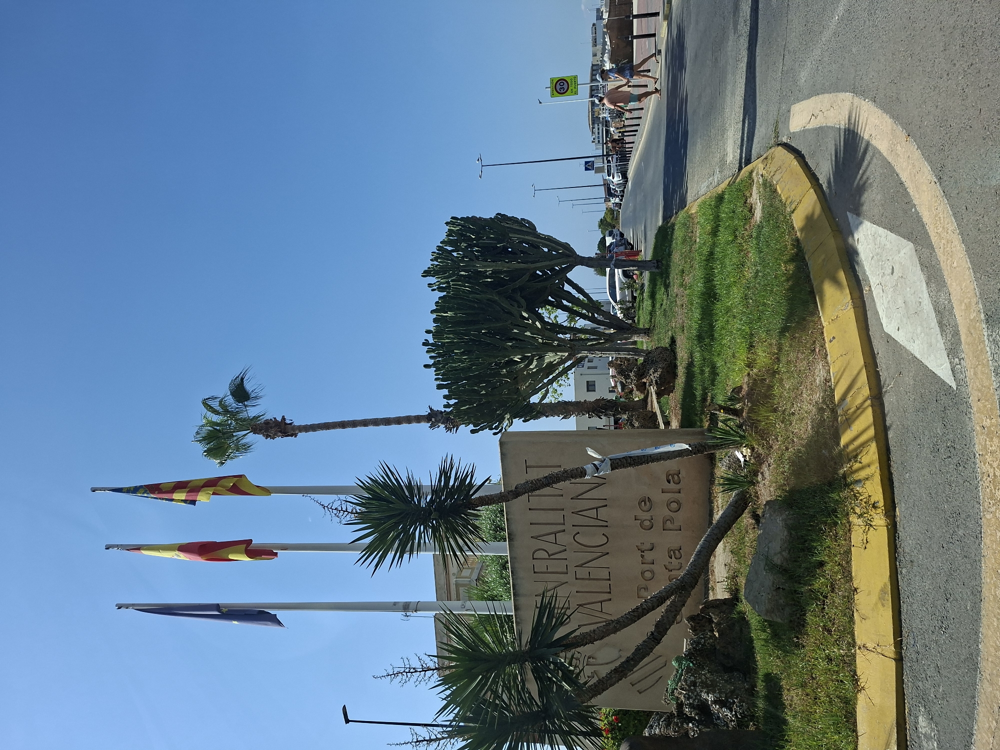 In Santa Pola, Spain, on a clear, sunny day, a concrete monument displays the text "GENERALITAT VALENCIANA Port de Santa Pola" carved into its surface. Three flags fly above it: the European Union flag on the left, the Spanish flag in the middle, and the Valencian Community flag on the right. To the left of the monument, Yucca plants and rocks are visible. To the right, a road curves away from the monument, bordered by a grassy area and trees, including a tall palm tree and a large Euphorbia tree. The road has traffic markings and a speed limit sign indicating 30. A few cars are parked along the side of the road, and two people, a man and a woman, are walking away from the camera on the right side of the road. The scene appears to be at the entrance to a port or public facility.