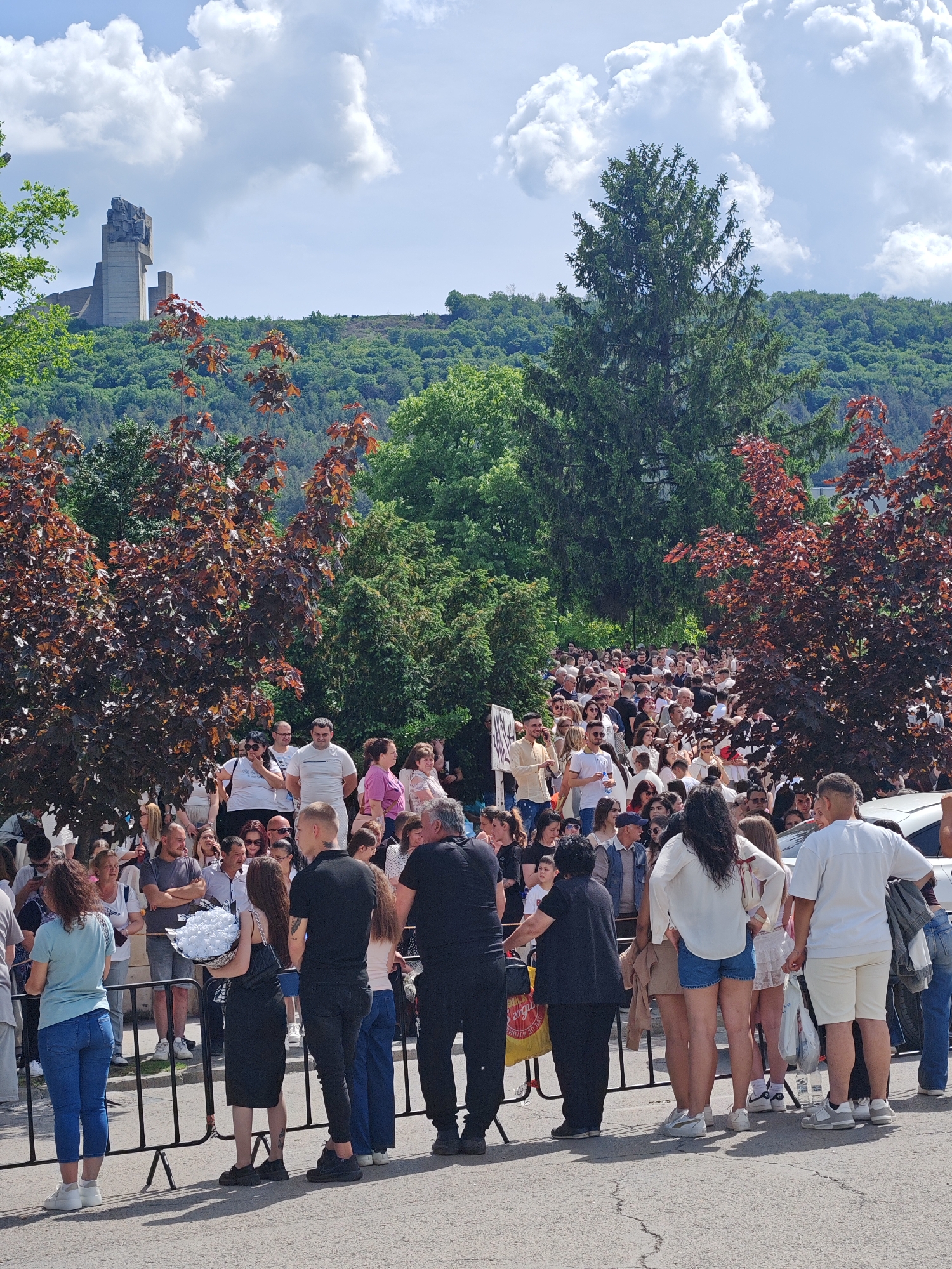 A large crowd of people are standing behind a black metal barrier on a paved surface. Most of the people in the crowd appear to be waiting, looking toward a point further up a path. The path leads up a slight incline through trees and foliage. In the background, atop a hill covered in trees, stands a large, grey, concrete monument. The location is Shumen, Bulgaria. The sky is partly cloudy. The day appears bright. Some of the people in the foreground are holding white flowers.