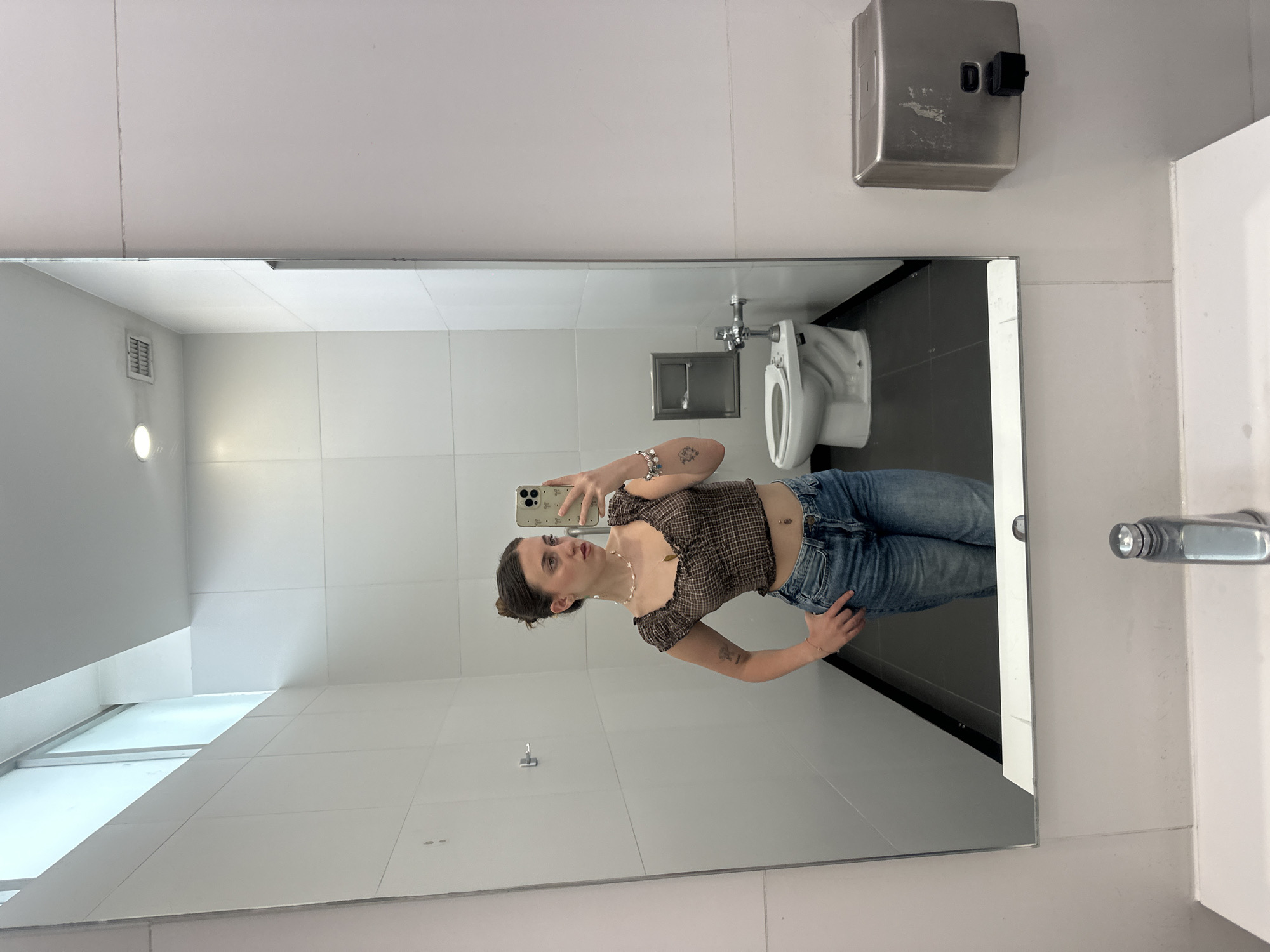 A woman is reflected in a mirror in a restroom. The restroom appears to be in Mexico City, Mexico. The woman is taking a photo of herself with a mobile phone. She is wearing a brown plaid shirt and blue jeans. A toilet is visible in the background. The restroom is clean and has white tiled walls. A soap dispenser is mounted on the wall above the mirror. Natural light is coming in from the window.