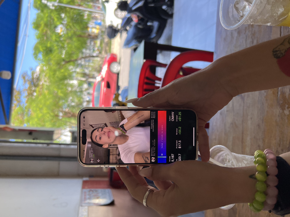 A person holds a smartphone showing a social media-like app interface. On the screen, a live video features a person posing with a peace sign. The app displays data, including "OMHV" symbol, a creator's revenue of +$1.25, and principal value of $17.5. There's also "Volume 5,167" and "Principal $23.7." The person holding the phone wears multiple beaded bracelets and a ring. Background shows outdoor scene with vehicles and trees.