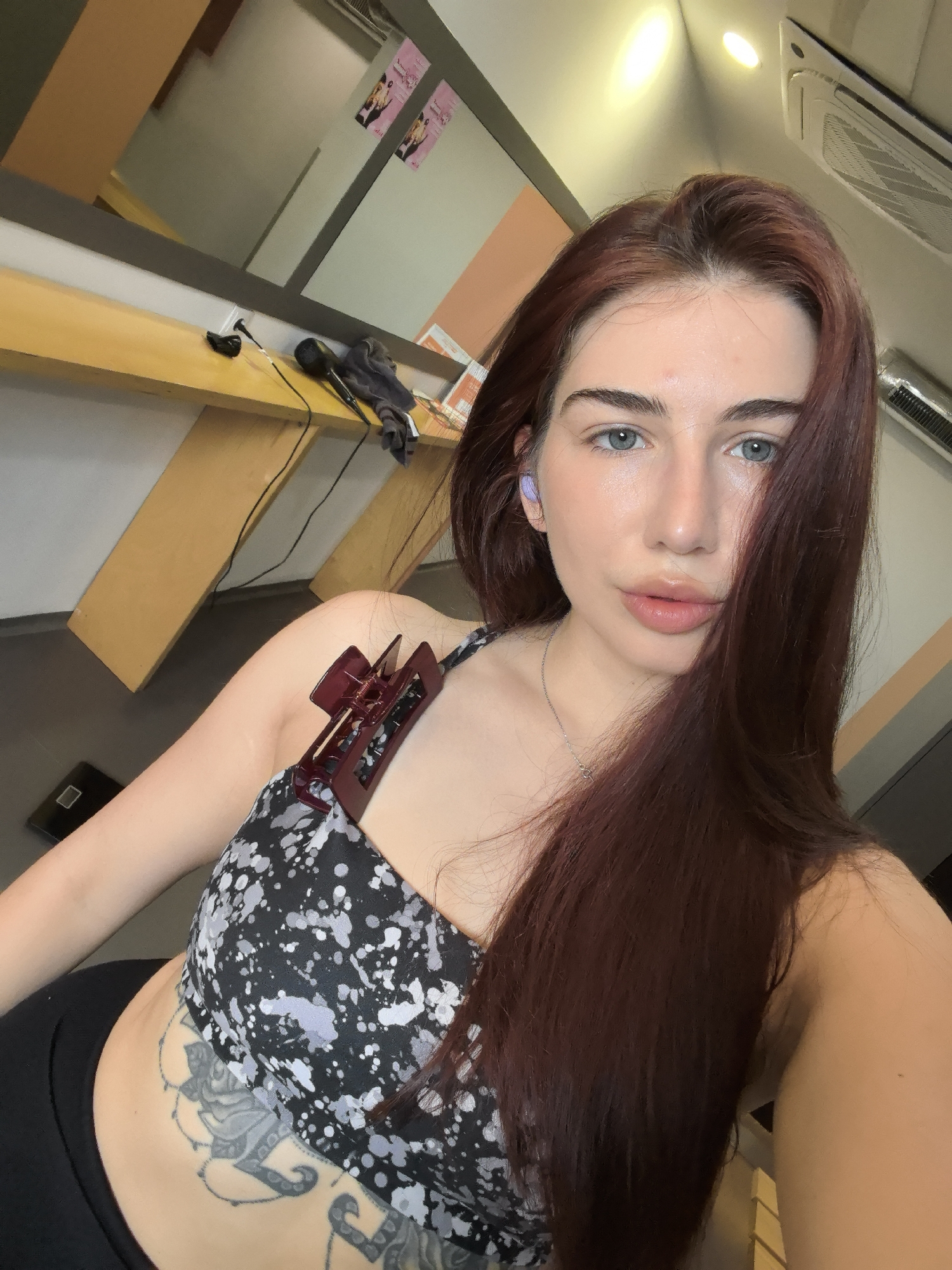 A young woman is posing for a selfie. She has long, dark red hair, blue eyes, and light skin. She is wearing a black and white sports bra and black leggings. A tattoo is visible on her abdomen. She has a maroon-colored hair clip attached to the strap of her sports bra. She also has a purple-colored earbud in her left ear.
The background suggests an indoor setting, possibly a salon or dressing room. There's a long wooden table against a wall with a mirror above it, along with a hairdryer. A small scale is visible on the floor in the lower left corner.
No specific details indicate the location is Stara Zagora, Bulgaria.