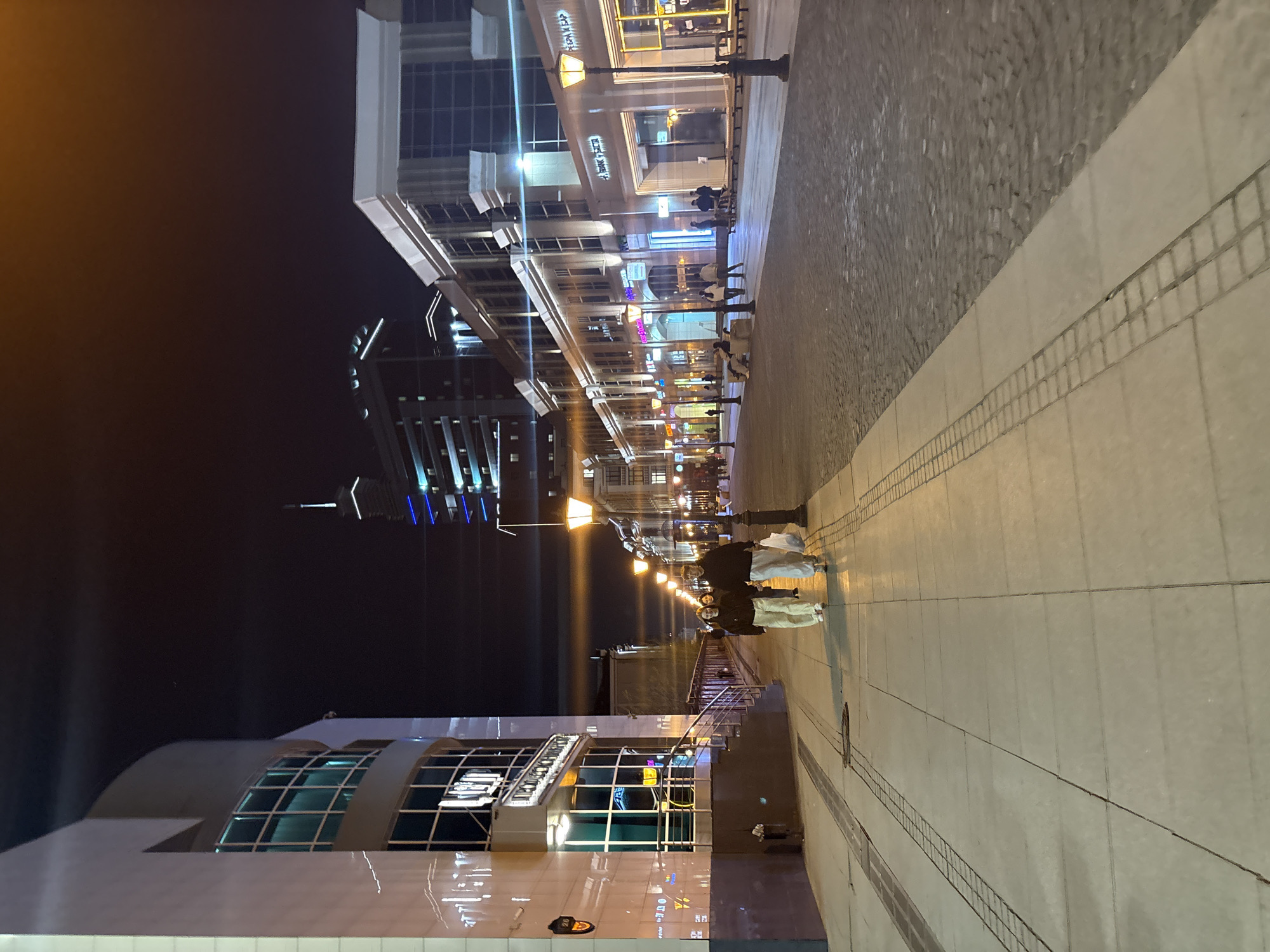 The image shows a nighttime scene on Bauman Street in Kazan, Russia. Buildings with lighted storefronts line the street, and streetlights illuminate the pedestrian walkway. People are walking along the sidewalk. The sky is dark.