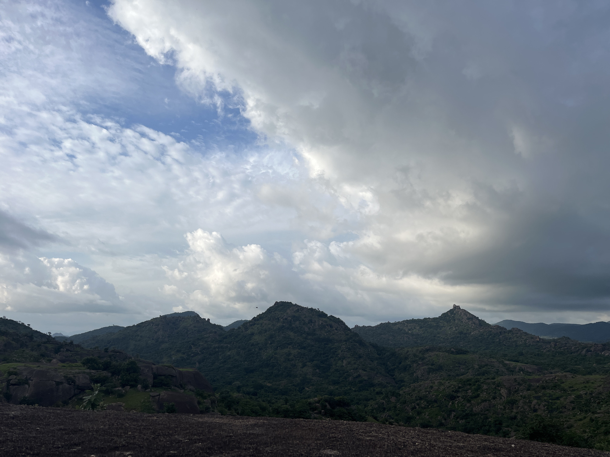 The image depicts a panoramic view of a mountainous landscape under a cloudy sky. In the foreground, a dark, rocky terrain is visible, with patches of dry vegetation. The middle ground is dominated by rolling hills covered in dense green vegetation and scattered large boulders. Several prominent peaks rise from these hills, some with rocky summits. The sky above is a dramatic mix of light blue patches and heavy, dark grey clouds, suggesting an overcast or approaching storm. The overall scene conveys a sense of rugged natural beauty and a remote, untamed environment. There are no people or man-made structures visible in the image. The location is described as Unknown, Unknown.