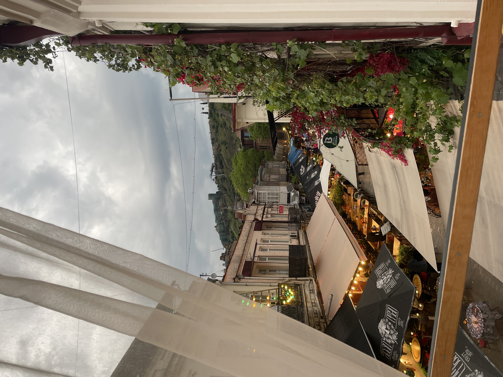 This image shows a view in Tbilisi, Georgia, possibly from a restaurant or cafe with outdoor seating. The sky is cloudy. The view looks down a street lined with buildings, with a hill and fortress visible in the background. The foreground features elements of a covered outdoor dining area. There are plants, including flowering vines, hanging from a structure above. Canopies and umbrellas provide shade to the dining area. The umbrellas are black and marked with advertising. There is some outdoor lighting. A partially visible white curtain is draped on the left side of the image.