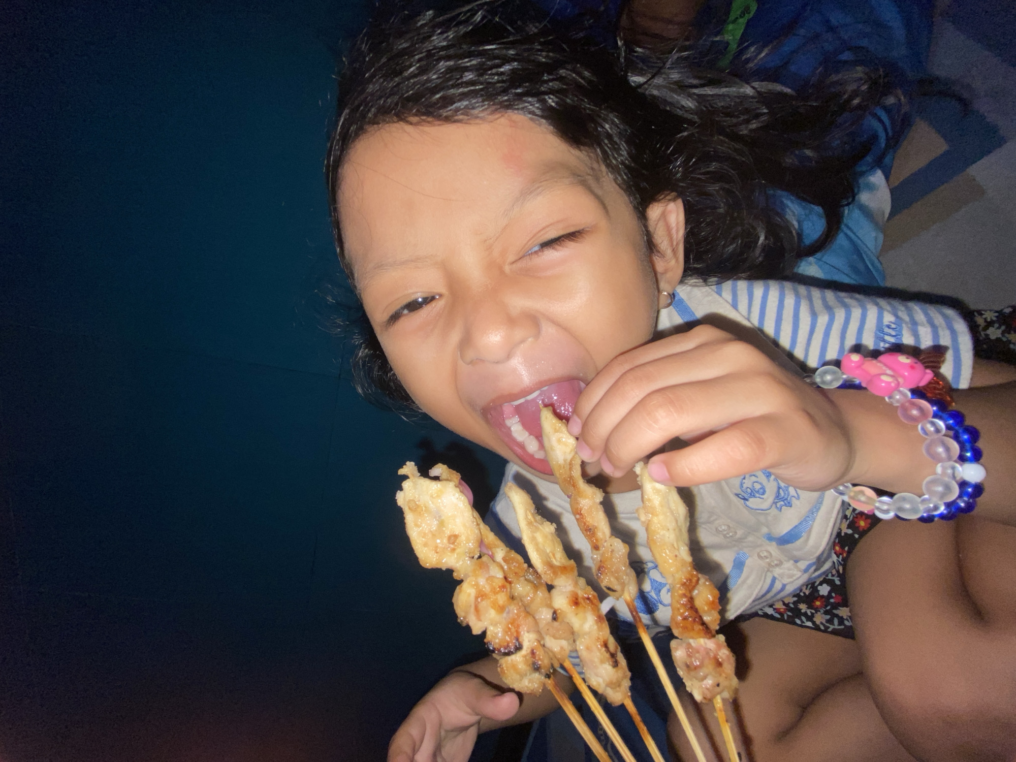 A young child with dark hair and an apparent bruise on their forehead is holding several skewers of grilled meat (likely chicken satay) and about to eat one. The child is wearing a striped shirt and a beaded bracelet. The location is most likely indoors, possibly a home, as the background is dark. The image does not provide sufficient information to confirm the location in Gelam, Indonesia.